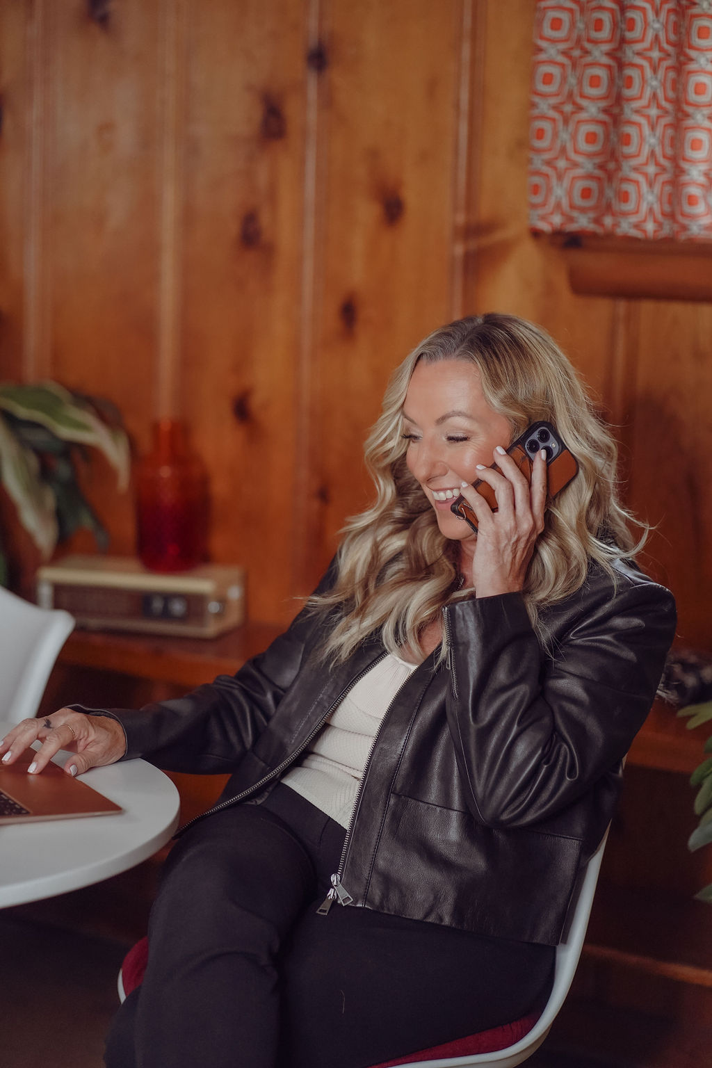 Woman with blonde hair talking on a phone, sitting at a table in a wood-paneled room.