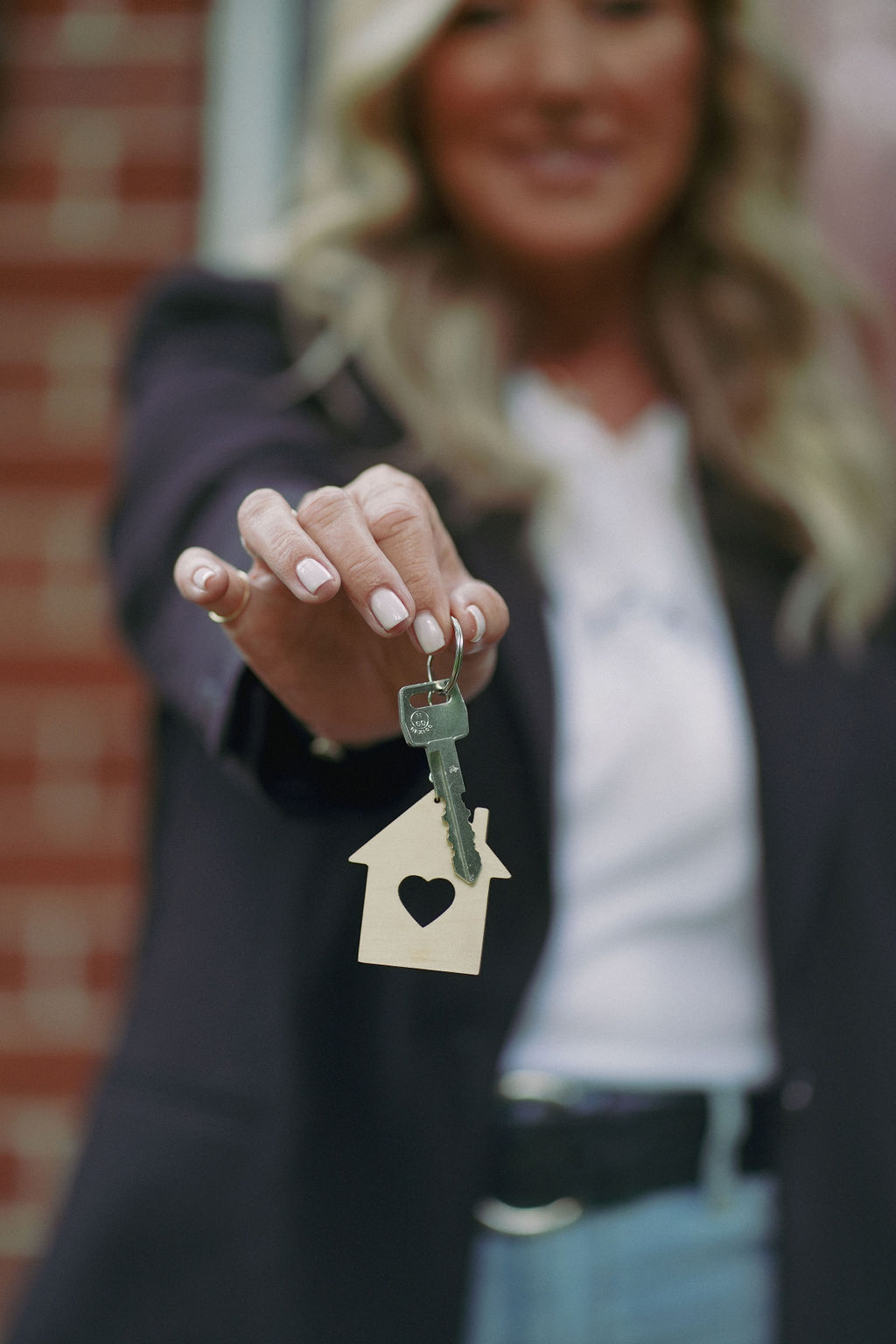 Woman holding a key with a house-shaped keychain, smiling, with a brick wall background.