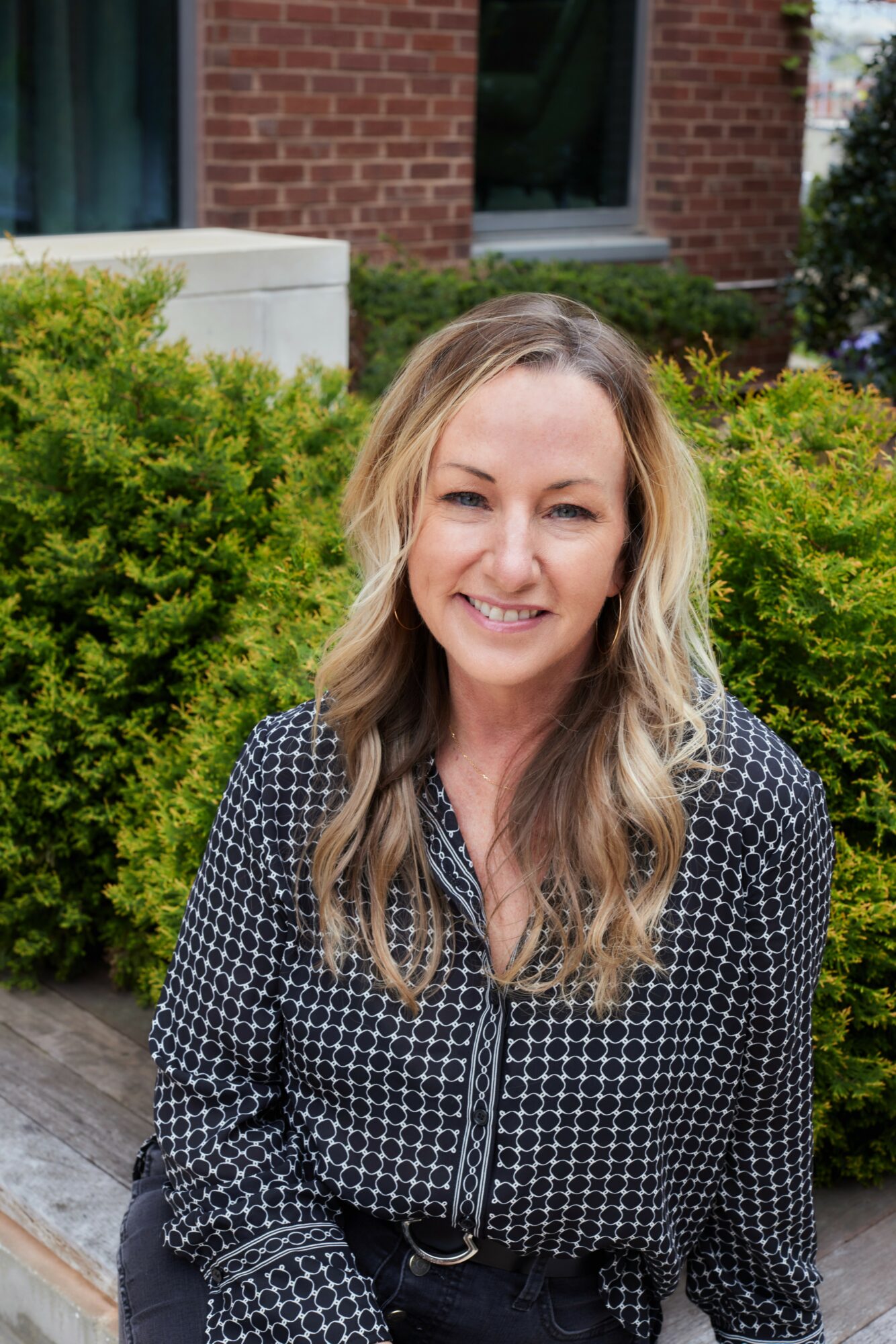 Smiling woman with long wavy hair standing outdoors in front of green bushes and a brick building, wearing a black and white patterned blouse.