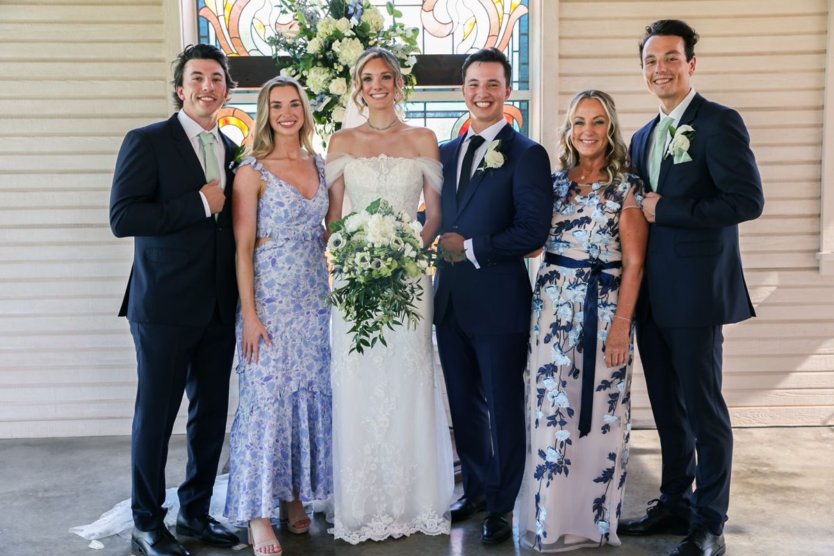 Group of six people in formal attire, standing indoors with a floral arrangement and stained glass window behind them.