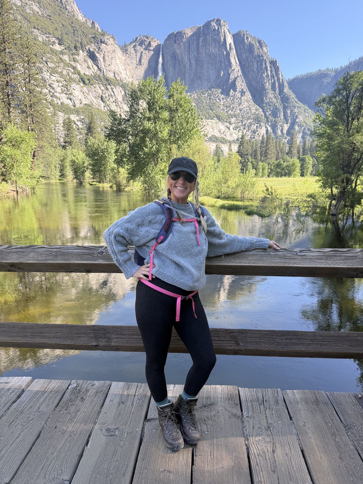 Woman standing on a wooden bridge with a river, trees, and mountains in the background, smiling, wearing a gray hoodie, black pants, and a cap.