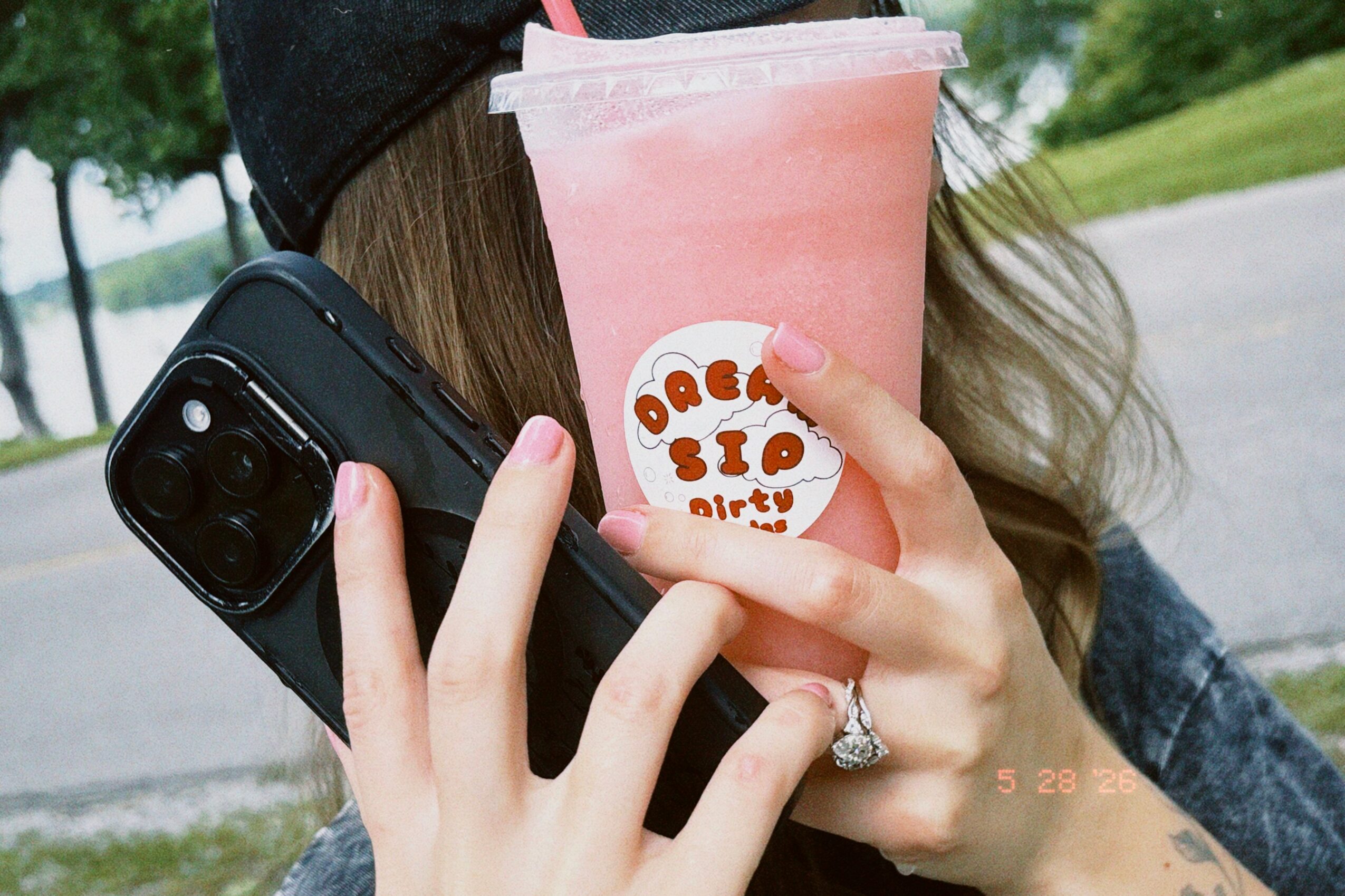 Person holding a pink drink, a phone, and wearing a ring outdoors with trees and grass in background.