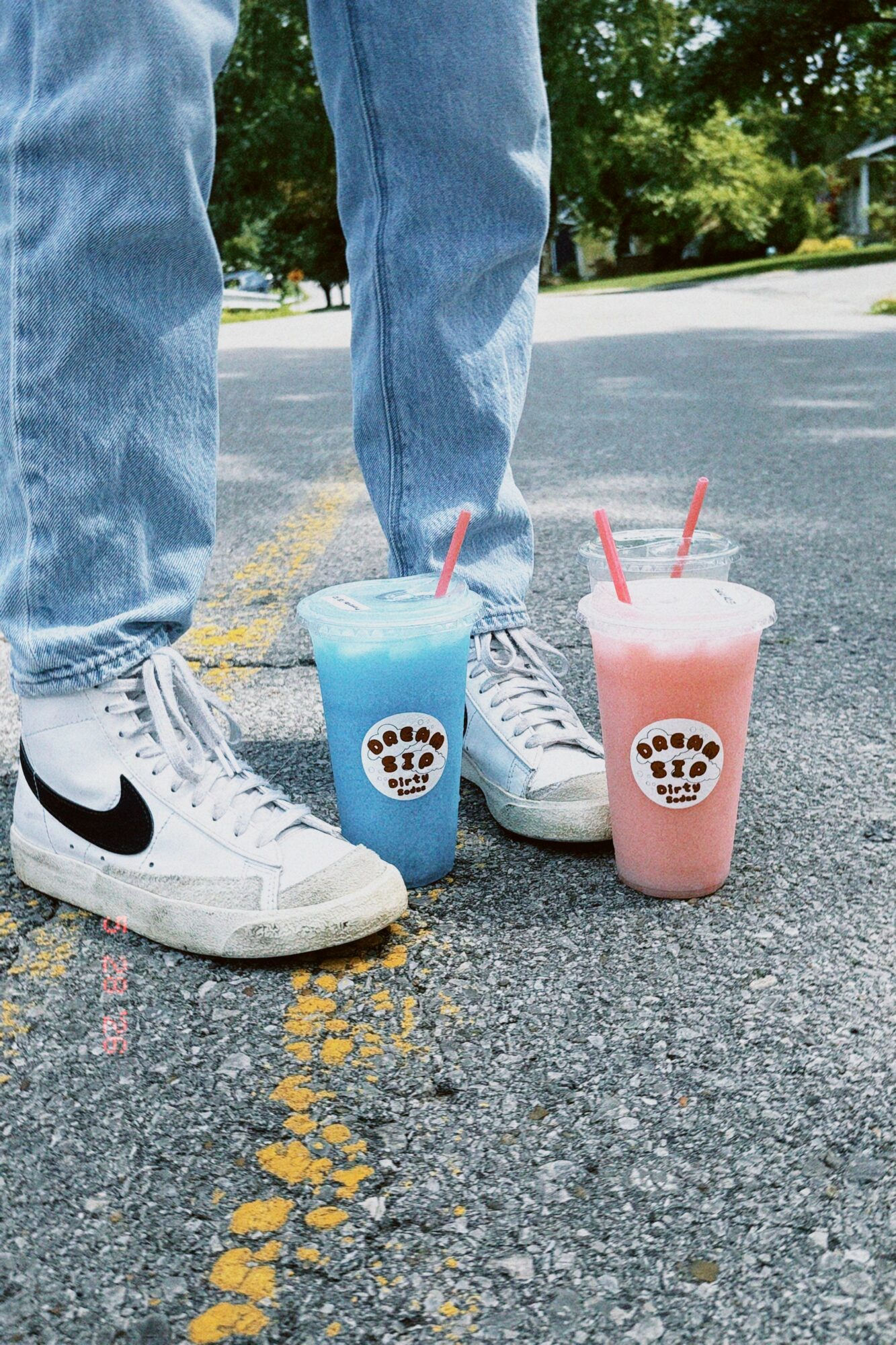 Person wearing white Nike sneakers and blue jeans standing on street with two colorful drinks in cups with straws.