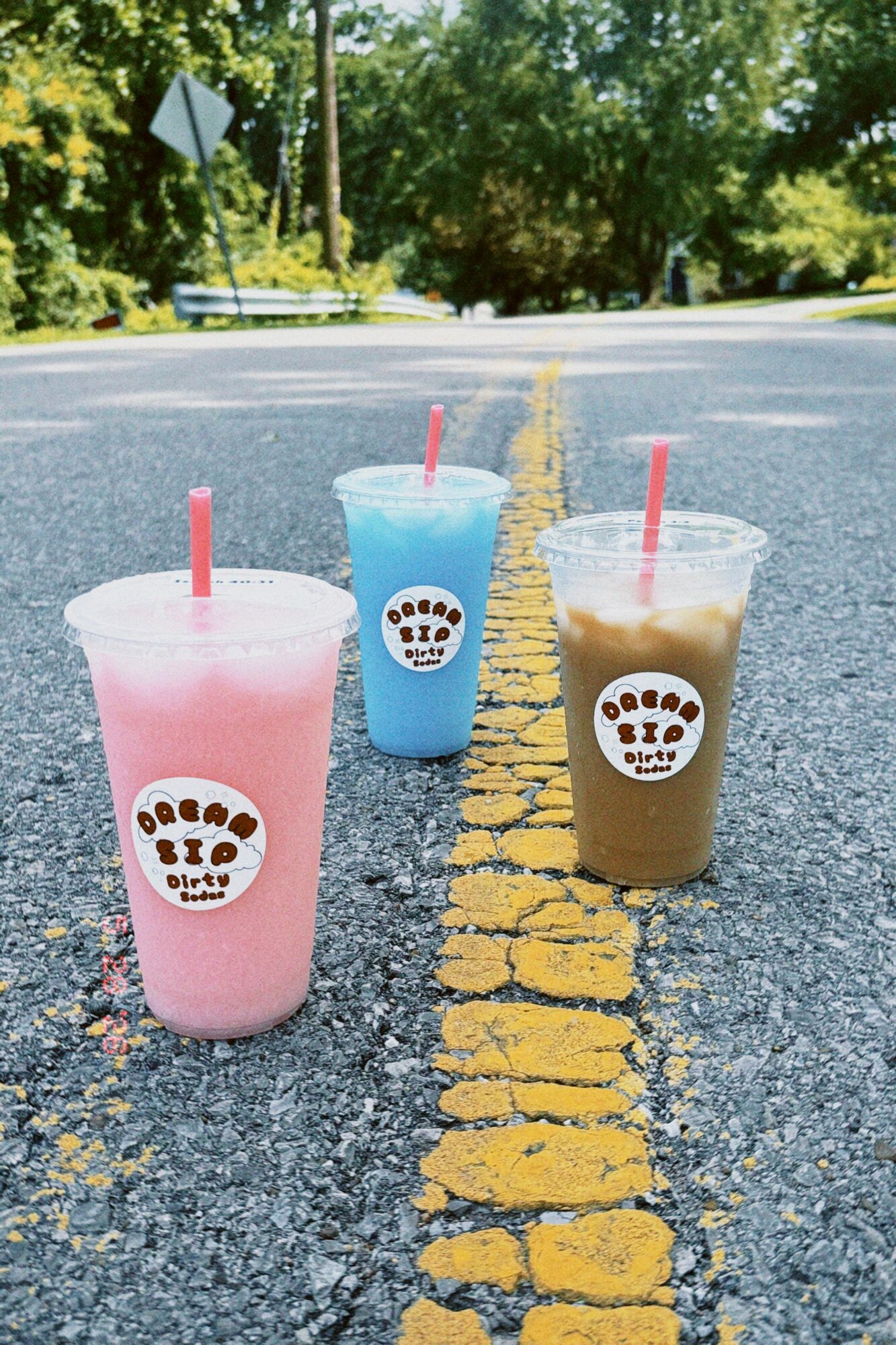 Three colorful smoothies with straws on a road with yellow lines, trees in background, sunny day.