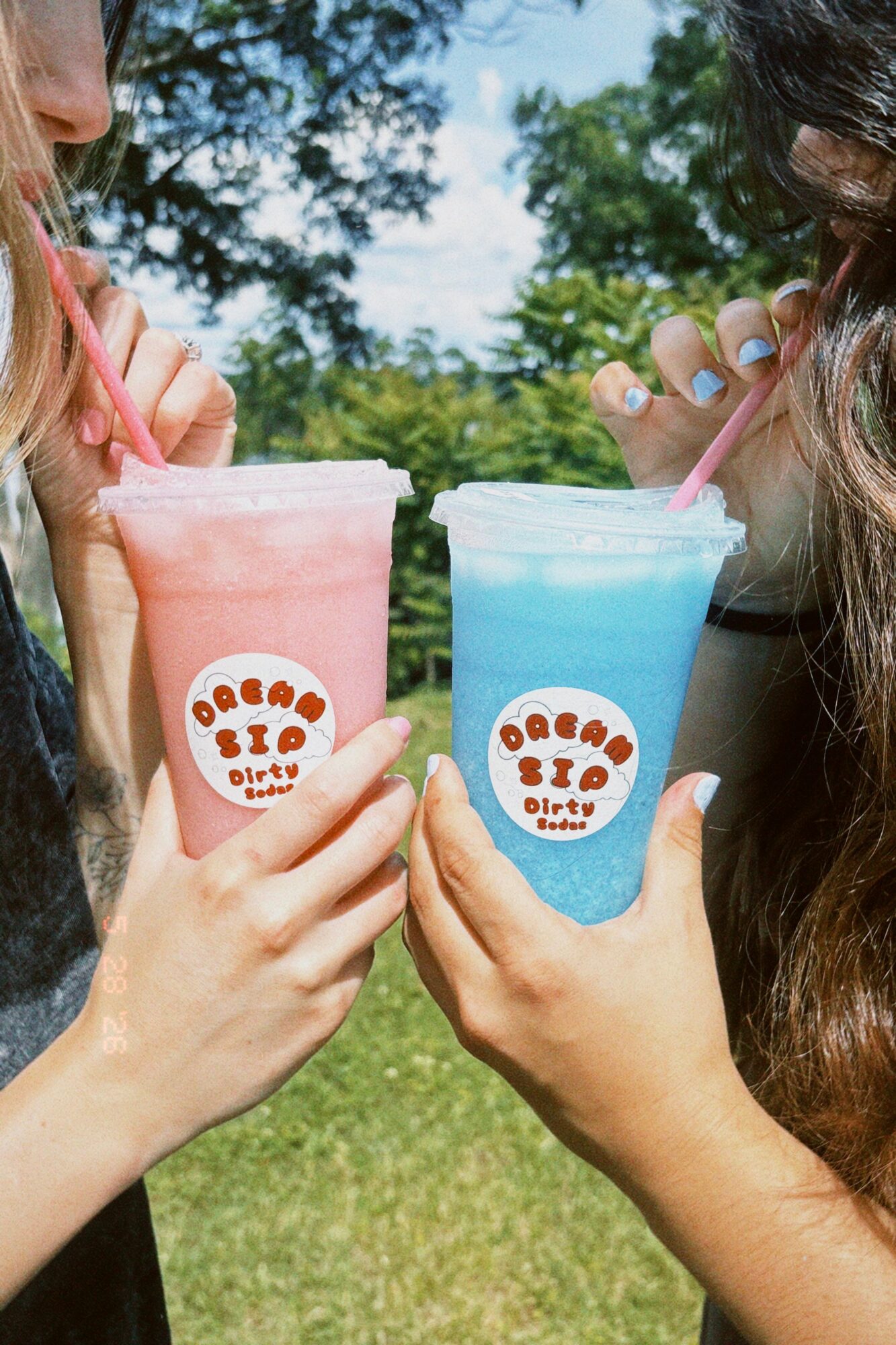 Two people holding colorful drinks with straws outdoors, trees and sky in background.