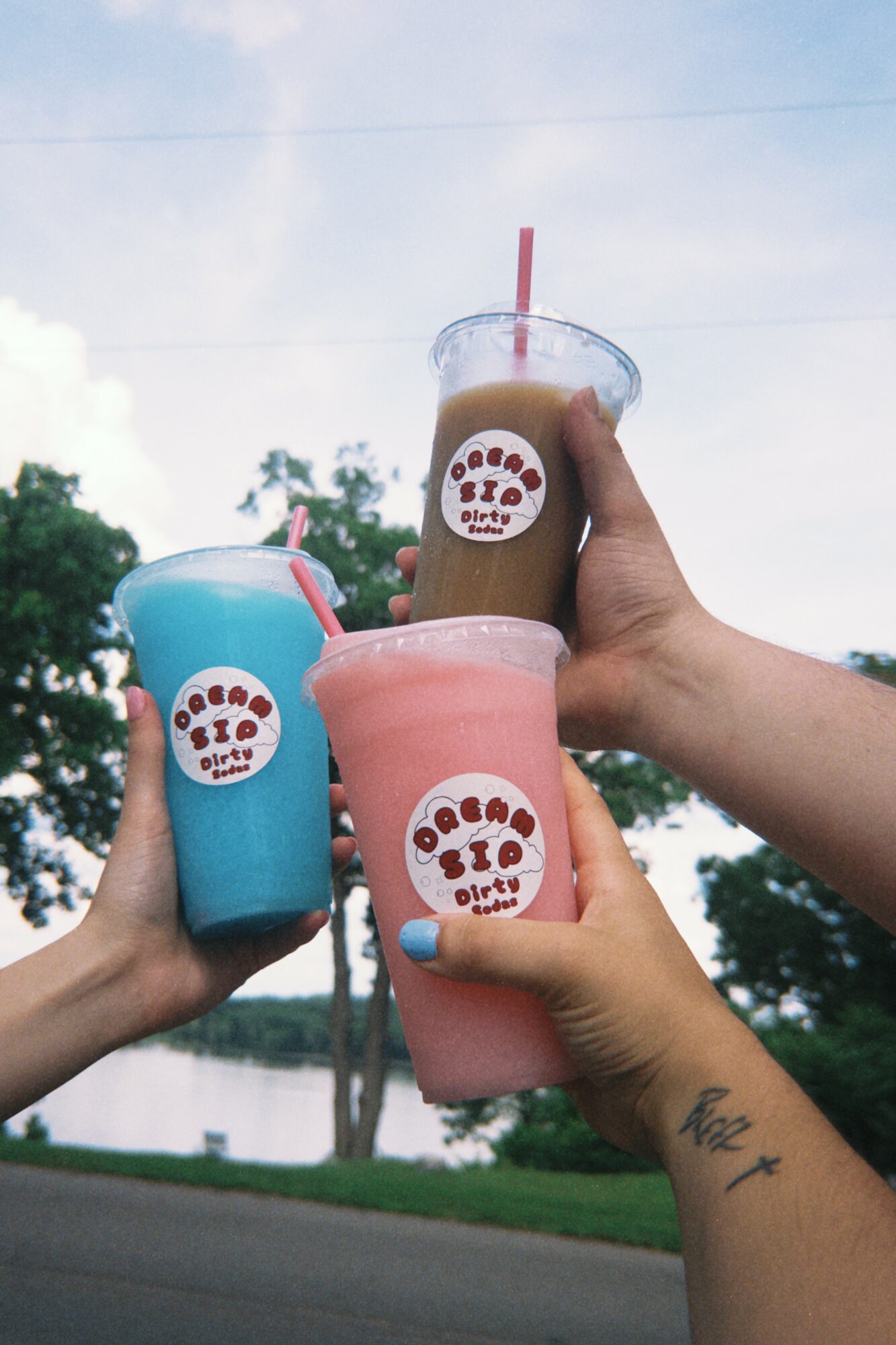 Three people holding colorful smoothies with straws outdoors, trees and sky in background.