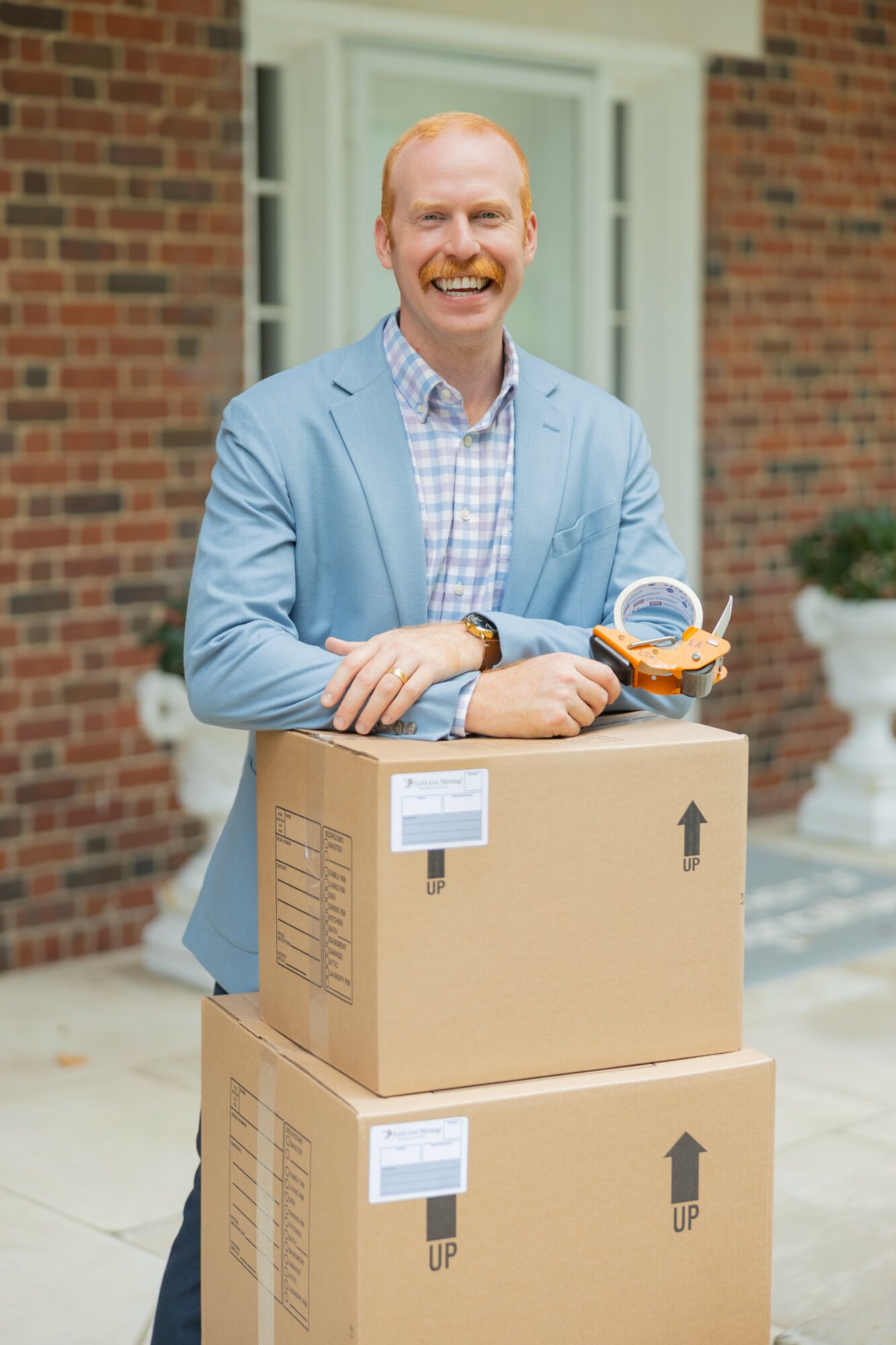Man with a big smile, wearing a light blue blazer and checkered shirt, standing outdoors with boxes and a clipboard.
