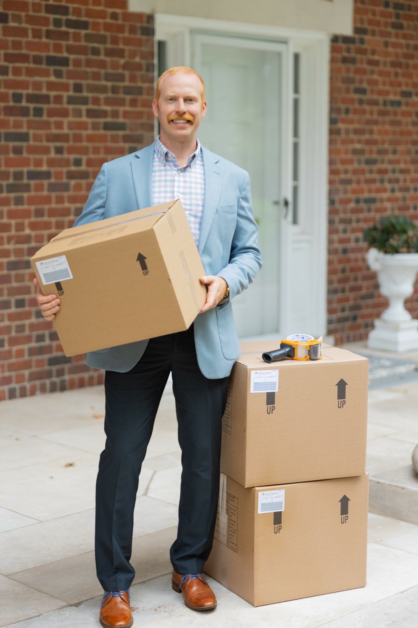 Man holding a large cardboard box outdoors in front of a brick building, with two smaller boxes on the ground beside him.