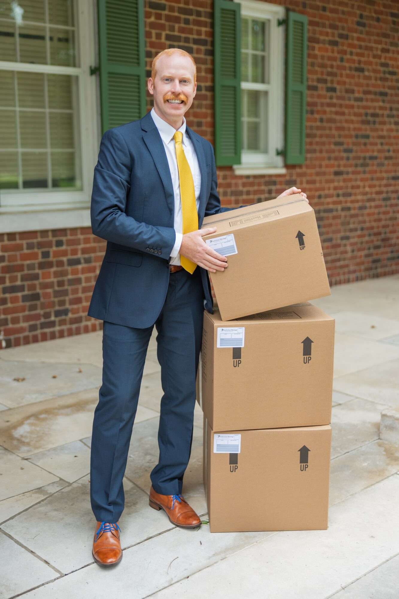 Man in suit holding a box, standing outside brick building with green window shutters.
