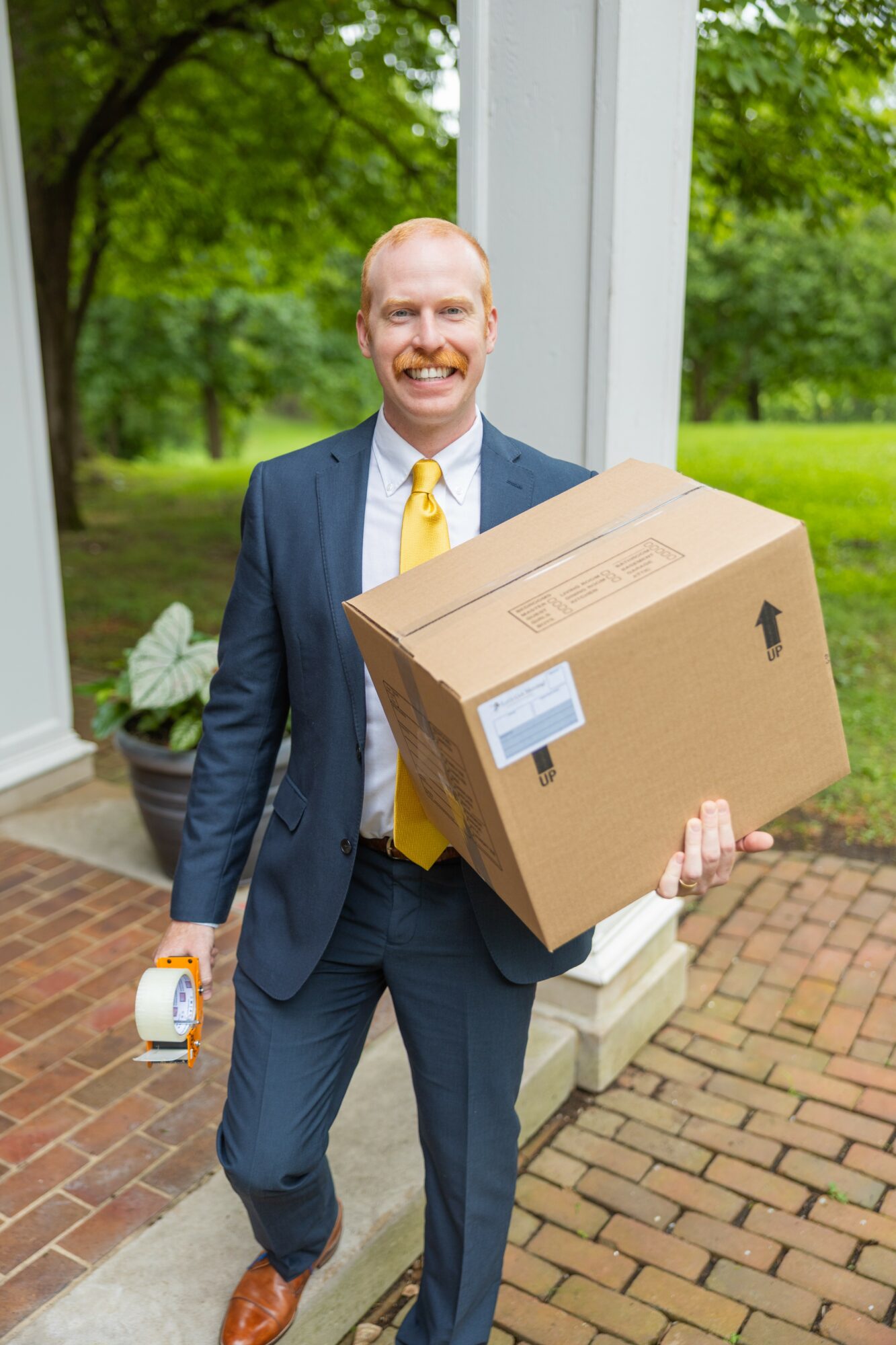 Man in suit holding a large cardboard box and a small item outside on a brick path, with trees and greenery in background.