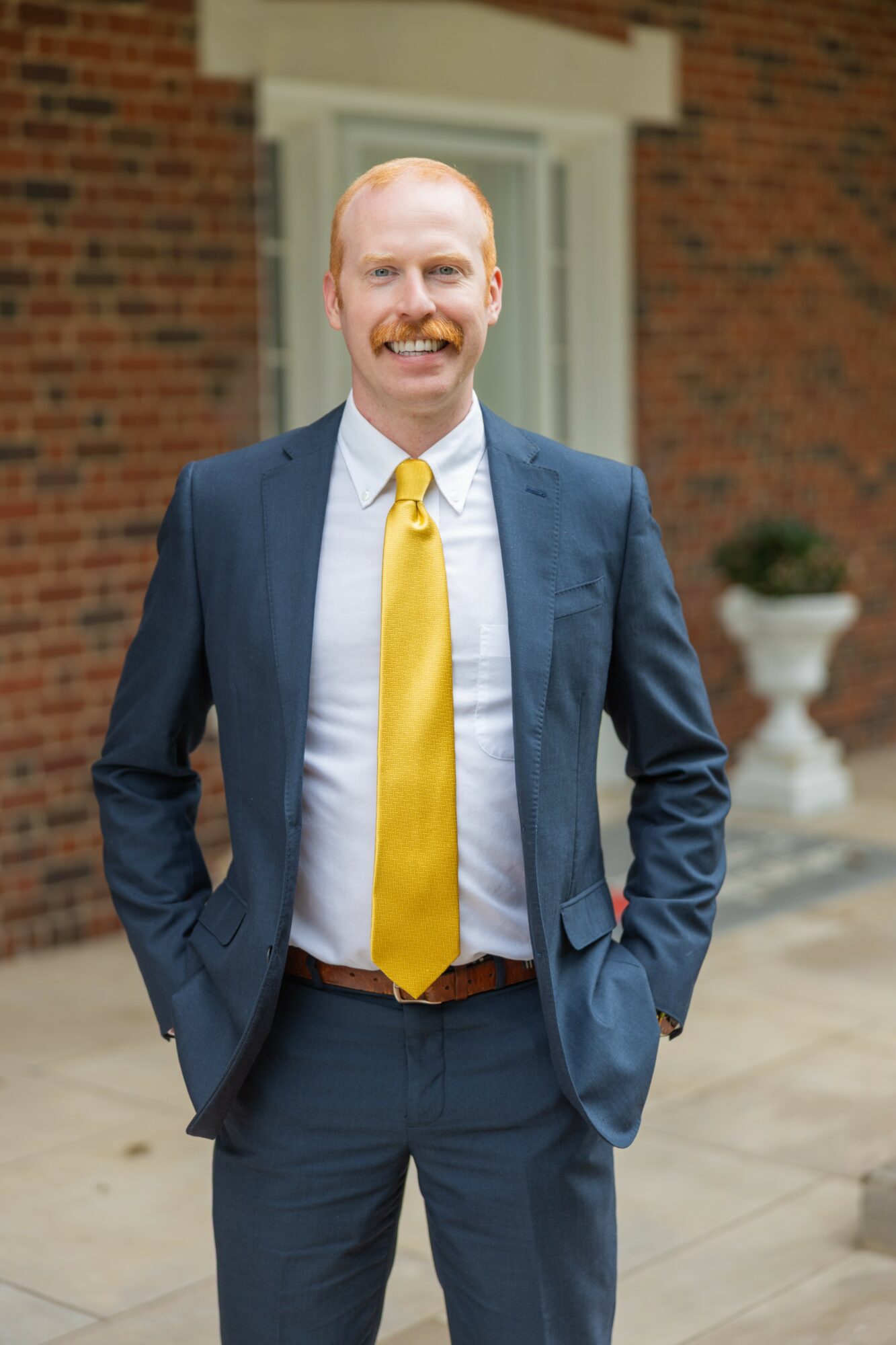 Man in a suit with a yellow tie standing outdoors in front of a brick building with potted plants.