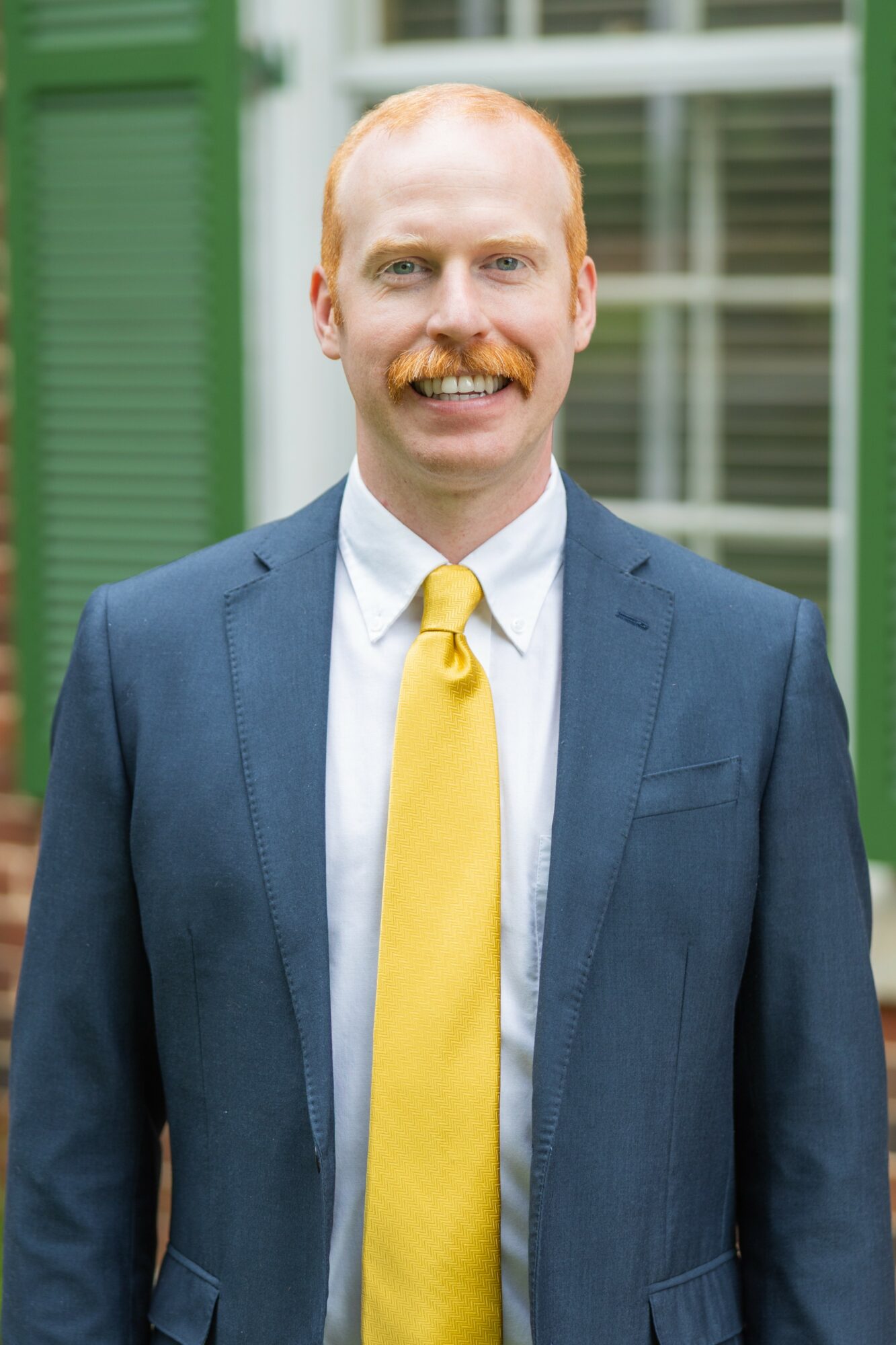 Man with red hair and mustache in a blue suit and yellow tie, smiling outdoors near a building with green shutters.