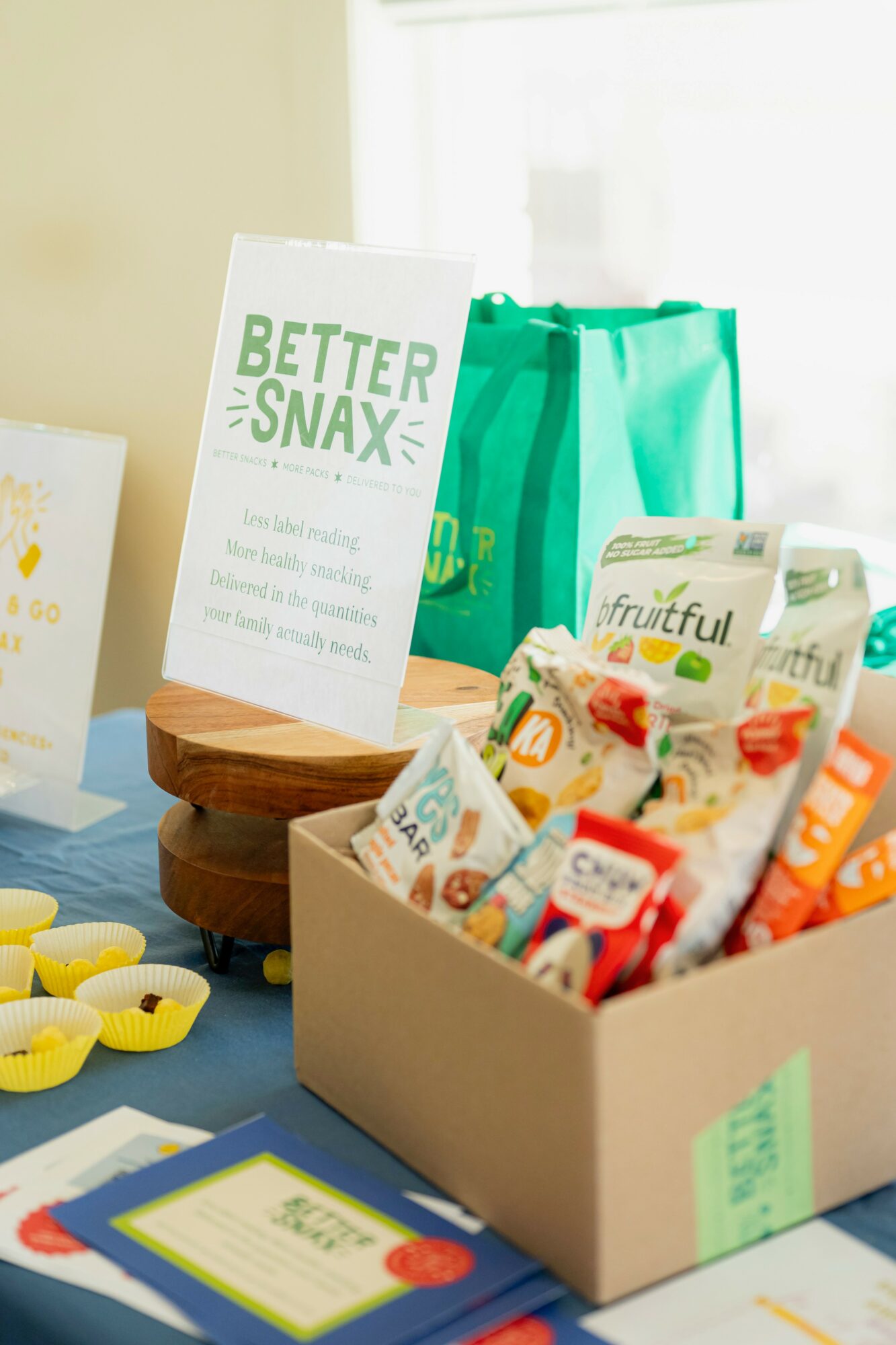 Open box of snack bars and candies on table with a green bag and a sign, bright room background.
