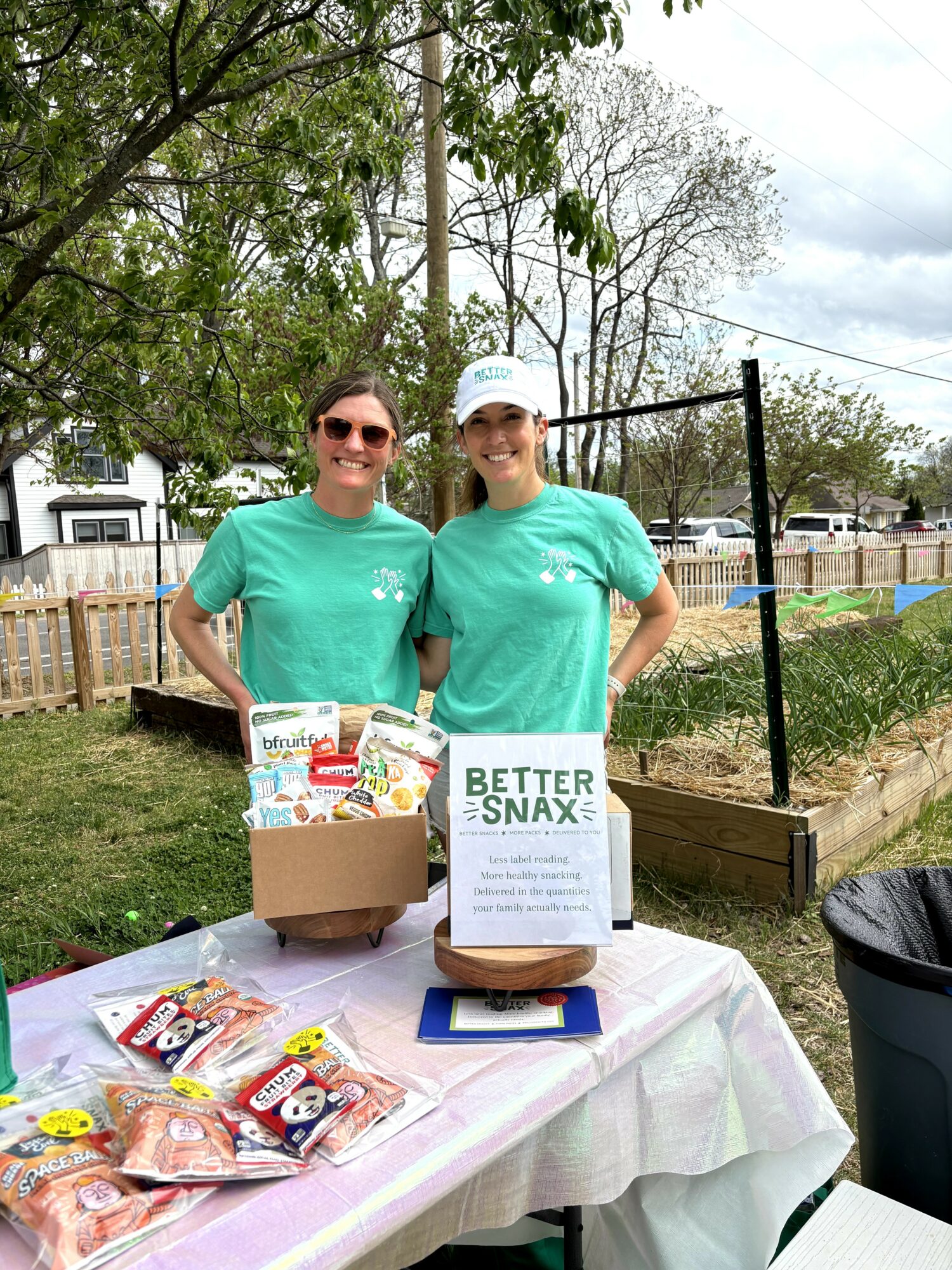Two women in teal shirts stand behind a table with snacks, outdoors with trees and a fence in background.