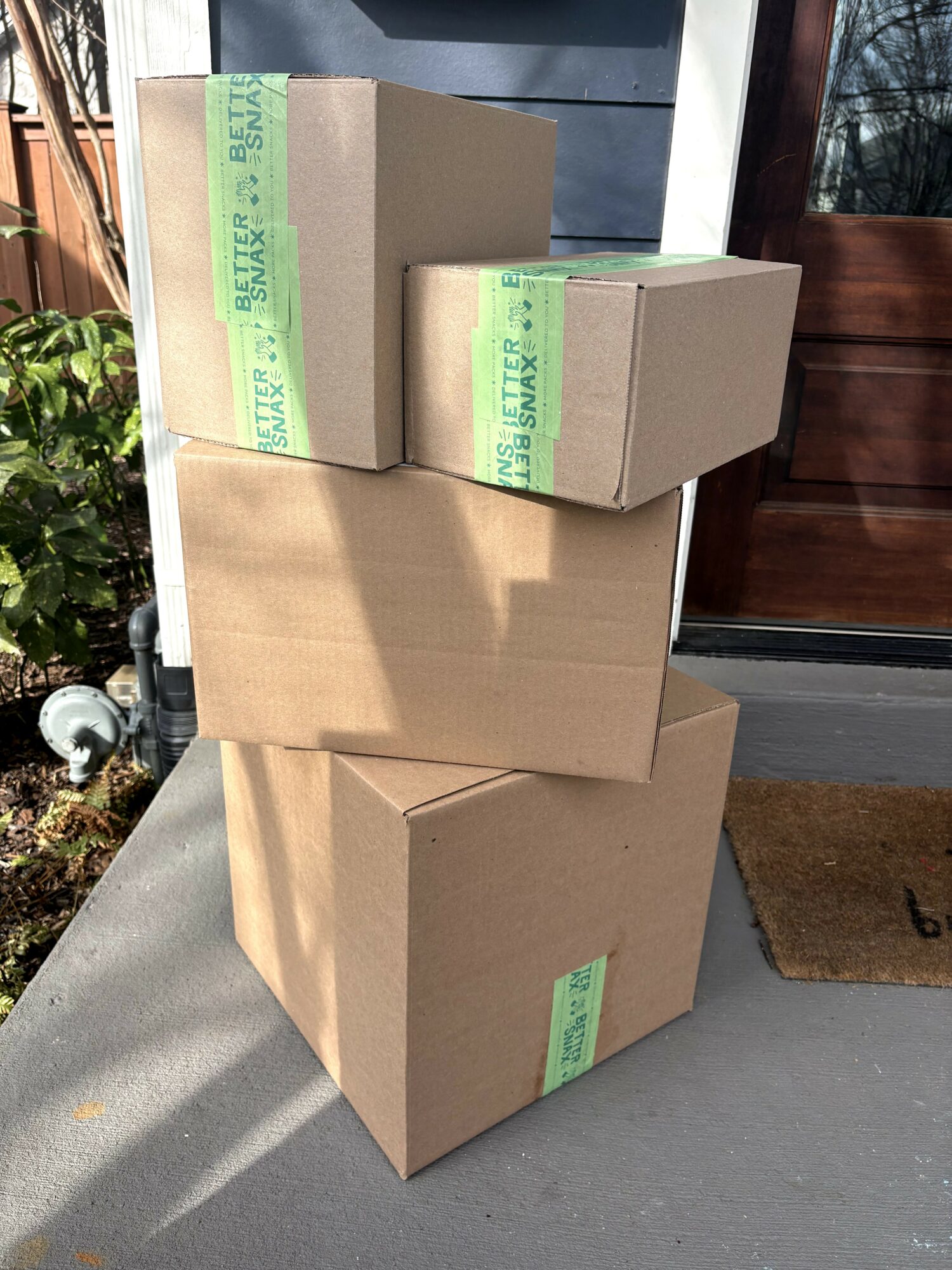 Four cardboard boxes stacked outside near a door, with sunlight and shadows, and a doormat nearby.