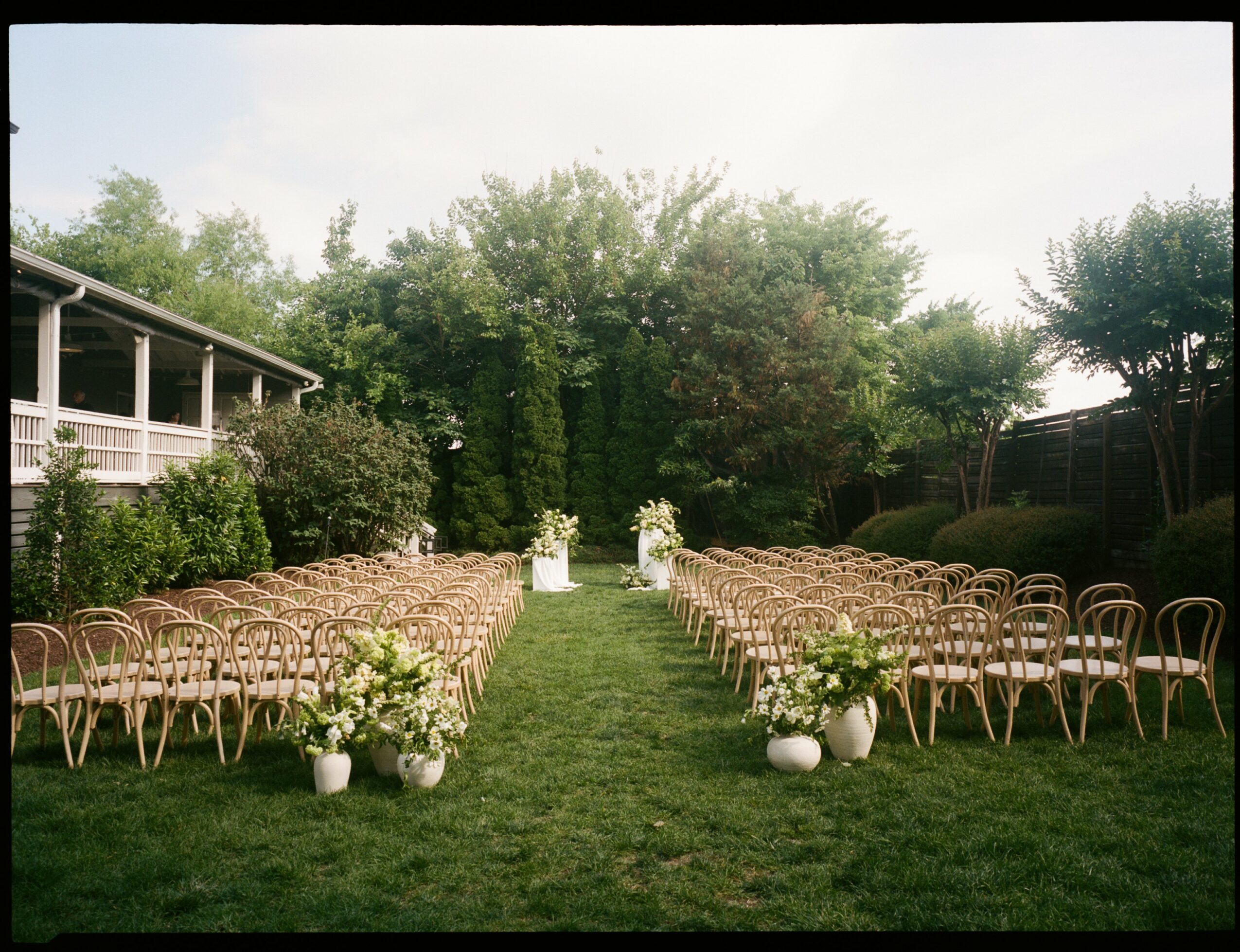 Outdoor wedding setup with chairs arranged on grass, decorated with white flowers, and an aisle leading to an arch.