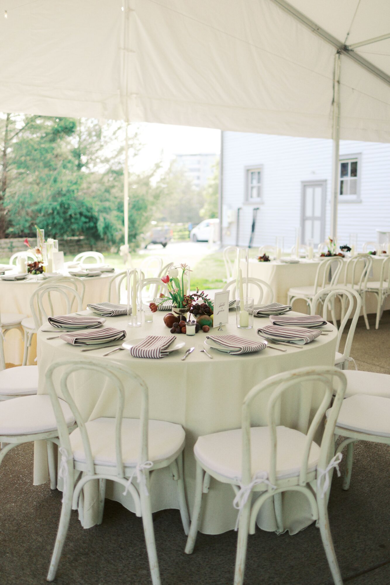 Round table with white chairs, set with plates, glasses, and a floral centerpiece, inside a tent with a view outside.