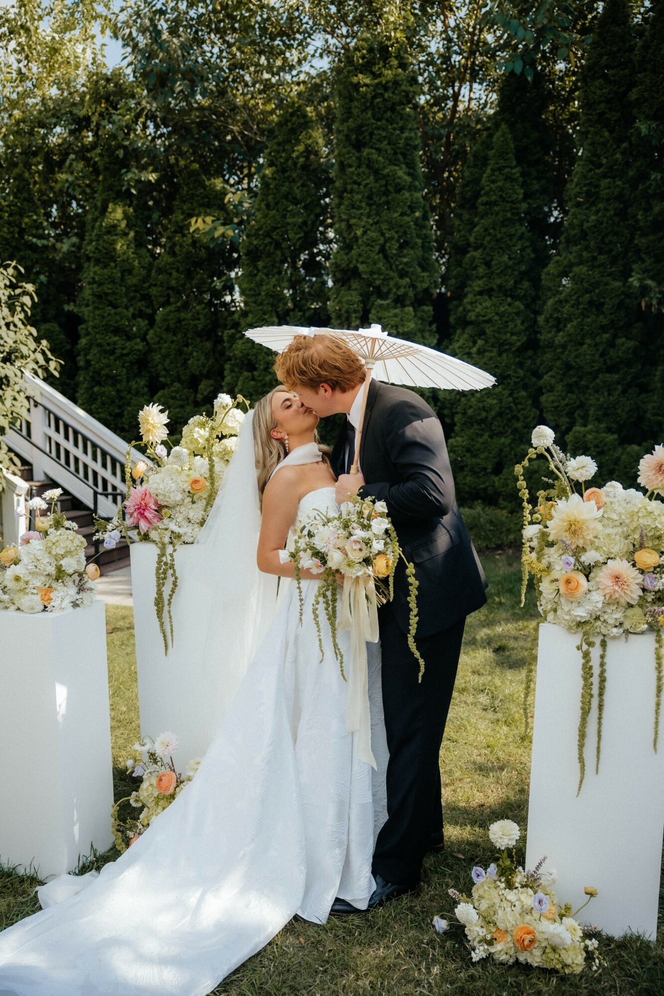 A bride and groom kiss outdoors under an umbrella, surrounded by floral arrangements and greenery.