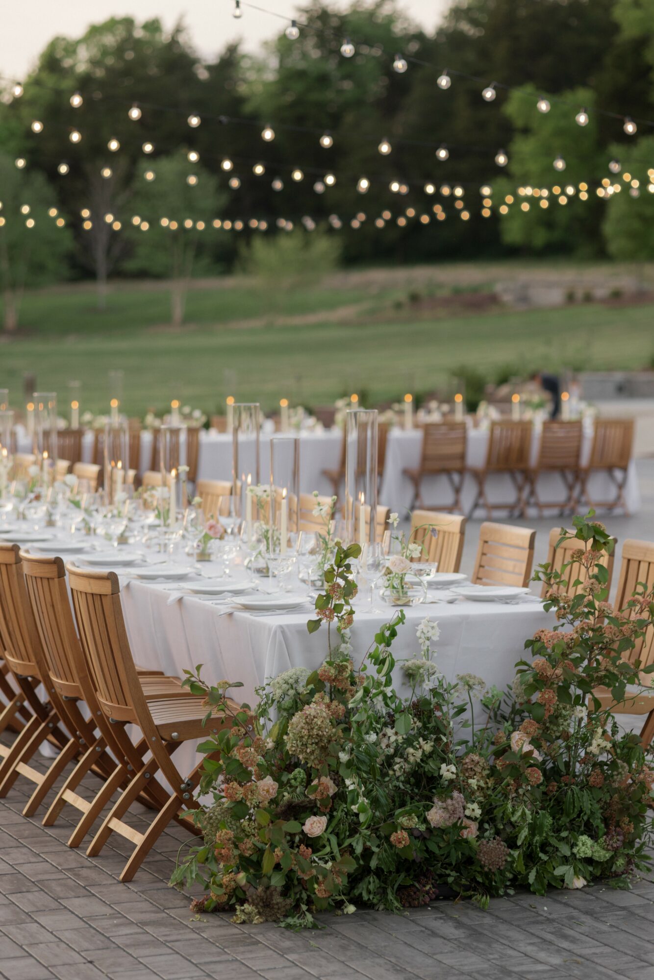 Long outdoor dining table with chairs, decorated with flowers and tableware, under string lights, with a grassy landscape background.