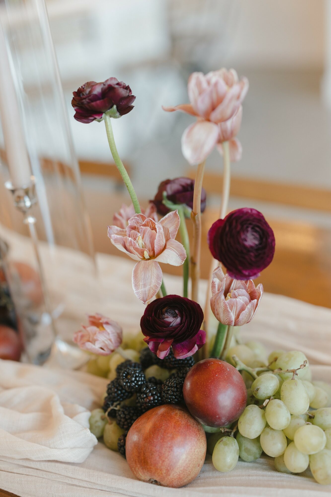 Arrangement of pink and dark purple flowers with apples and grapes on a table, blurred background.