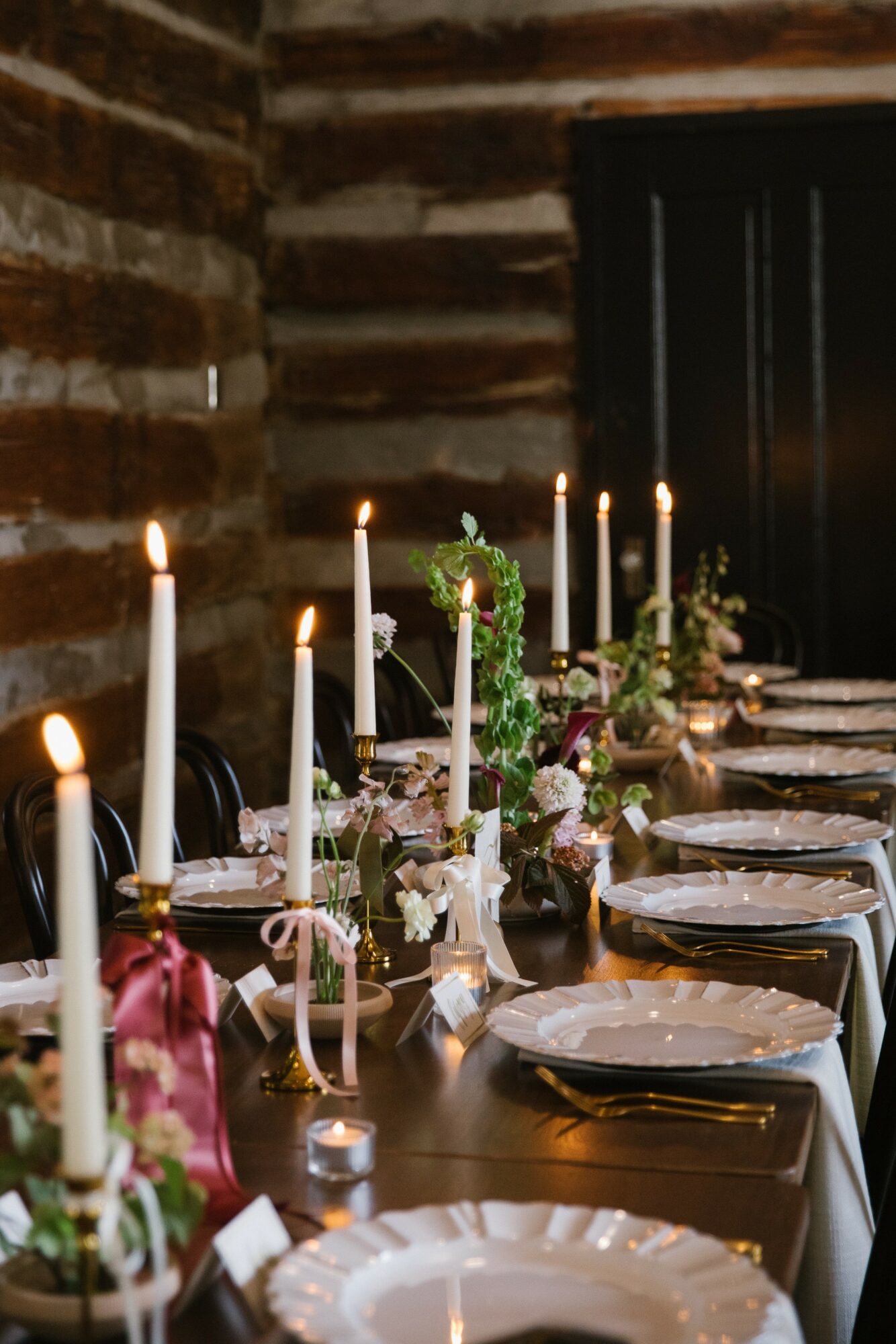 A long dining table set with white plates, candles, and floral centerpieces in a rustic brick room.