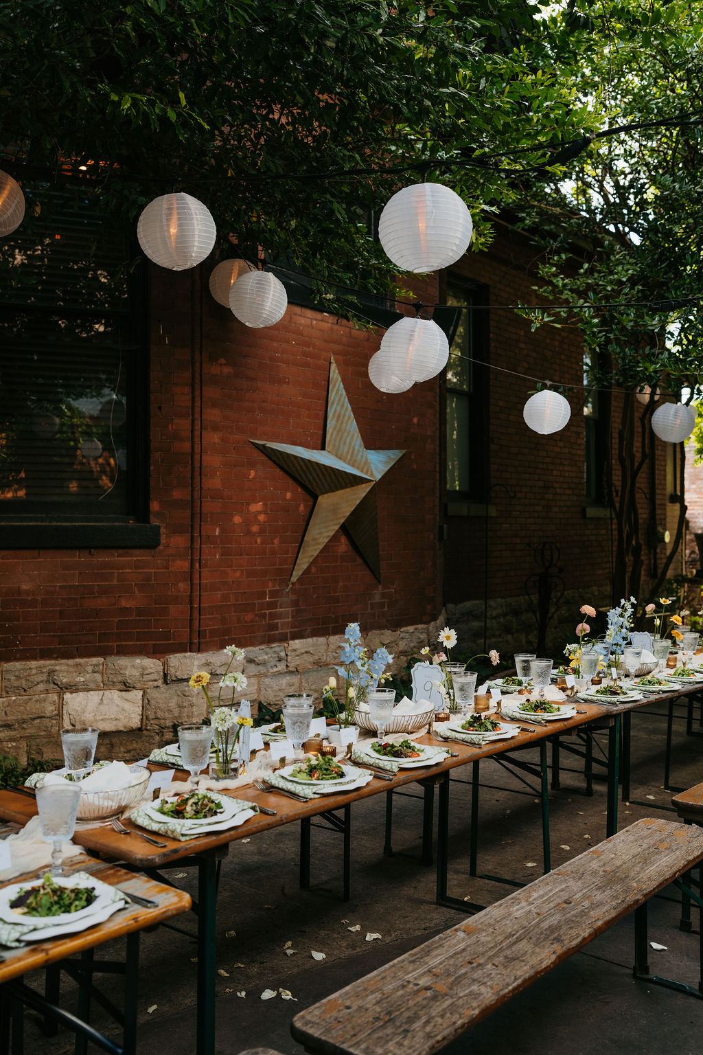 Outdoor dining setup with a long table, chairs, and hanging paper lanterns against a brick building with a star decoration.