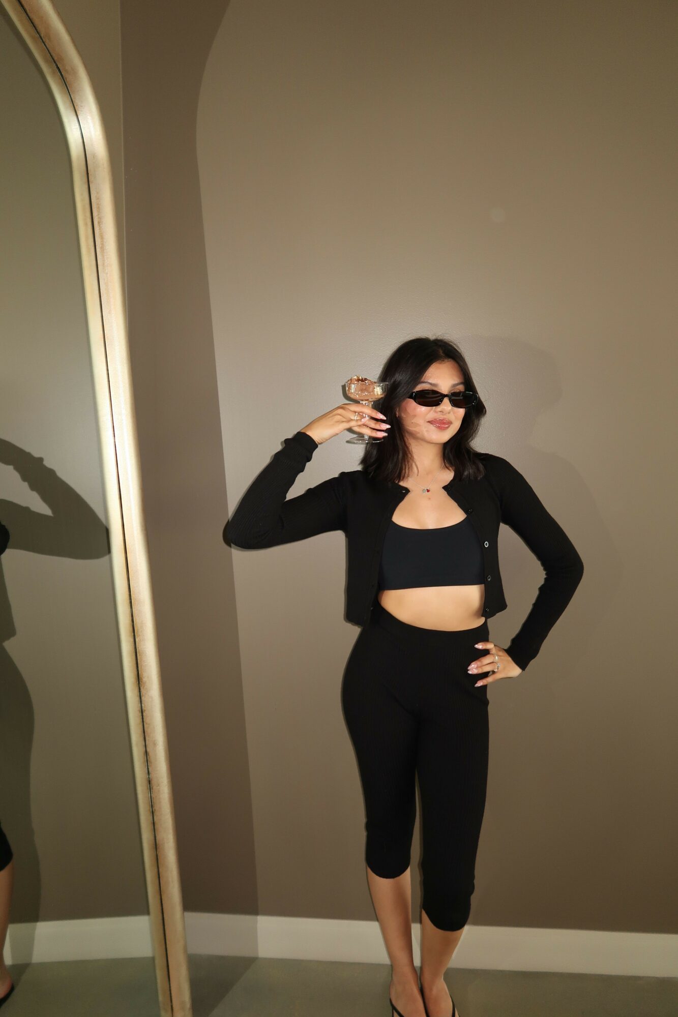 Woman in black outfit taking a mirror selfie, standing against a plain wall, holding phone with one hand, shadow visible on wall.