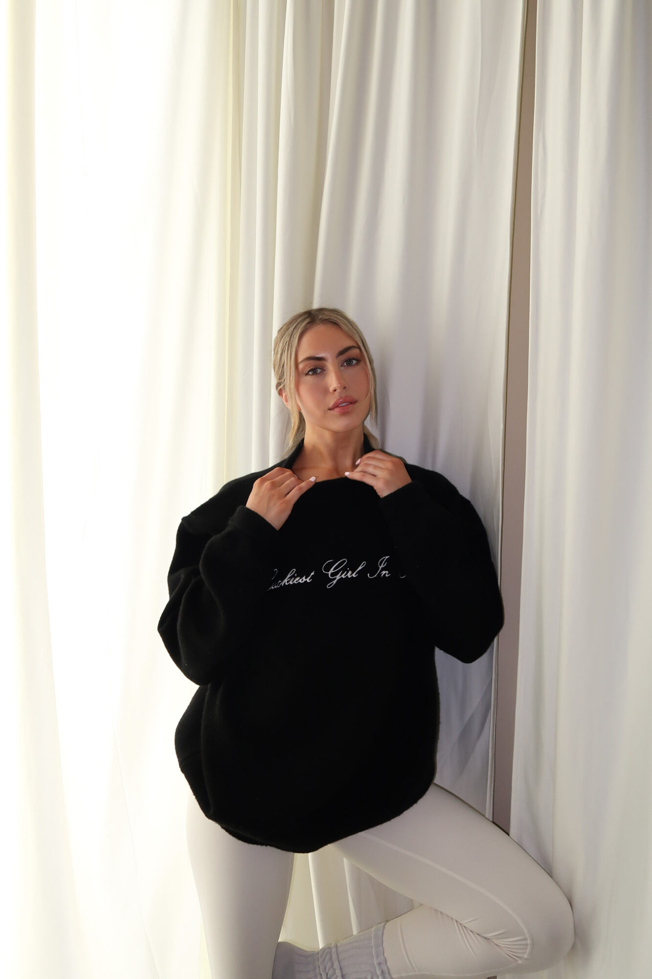 Woman with blonde hair in a black sweatshirt and white pants standing in front of white curtains.