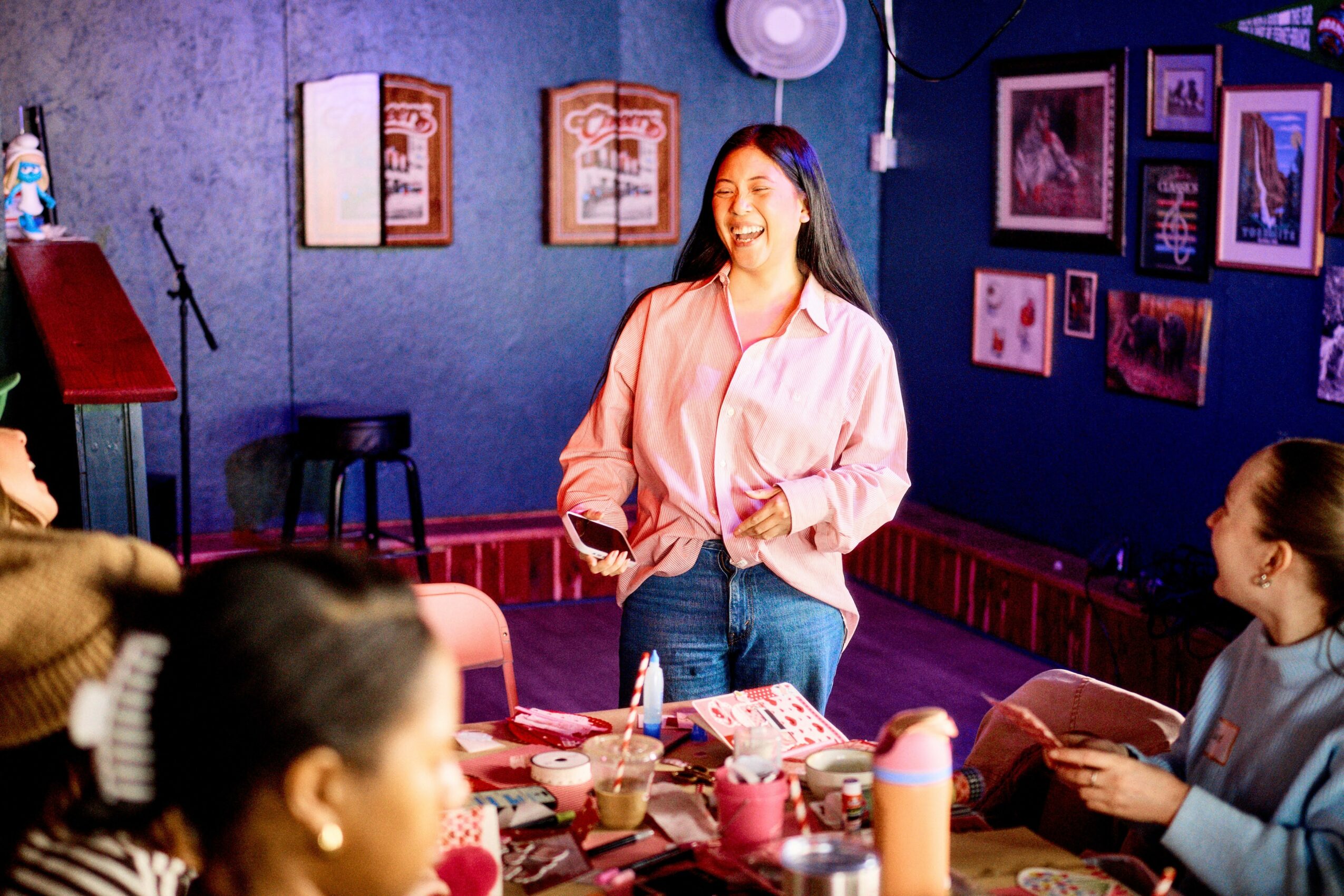 Woman standing and smiling in a room with people sitting at a table, colorful artwork on walls, and a microphone stand.