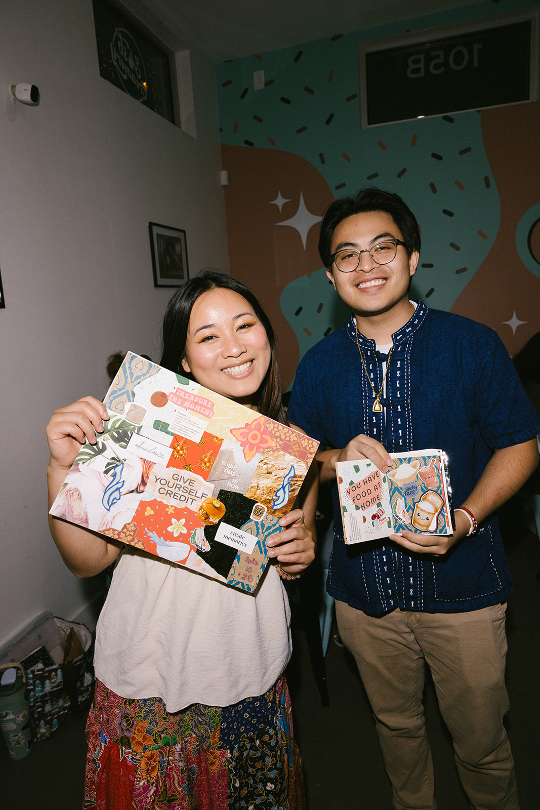 Two smiling people holding decorated photo frames indoors, with a colorful wall and framed pictures in the background.