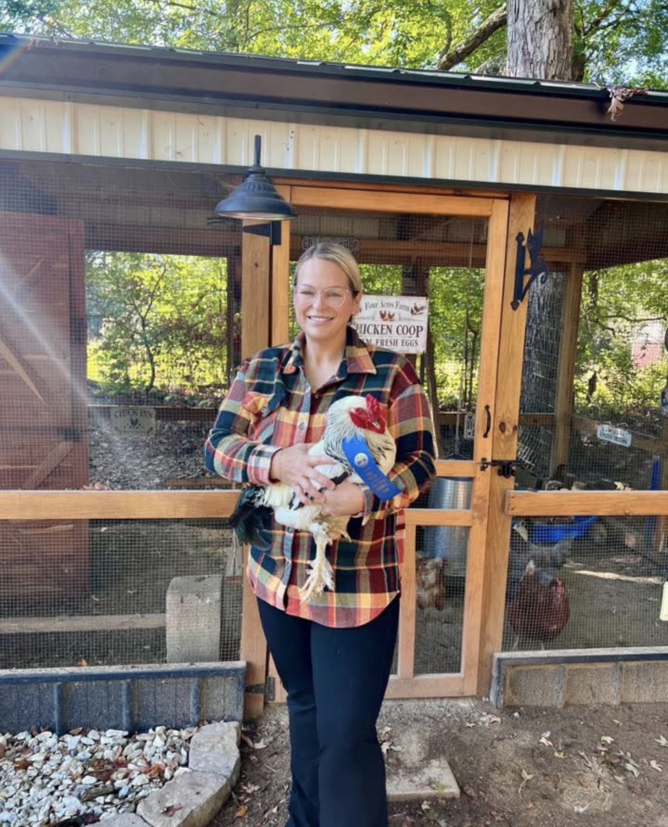 Woman holding a chicken and a blue toy, smiling outdoors near a wooden structure, trees in background.