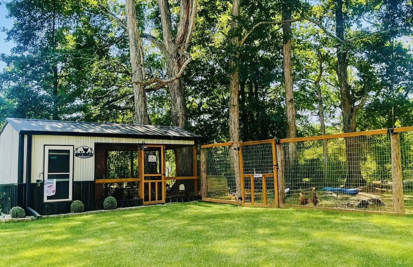 Small building with a porch next to a fenced outdoor area with animals, surrounded by trees and grass.