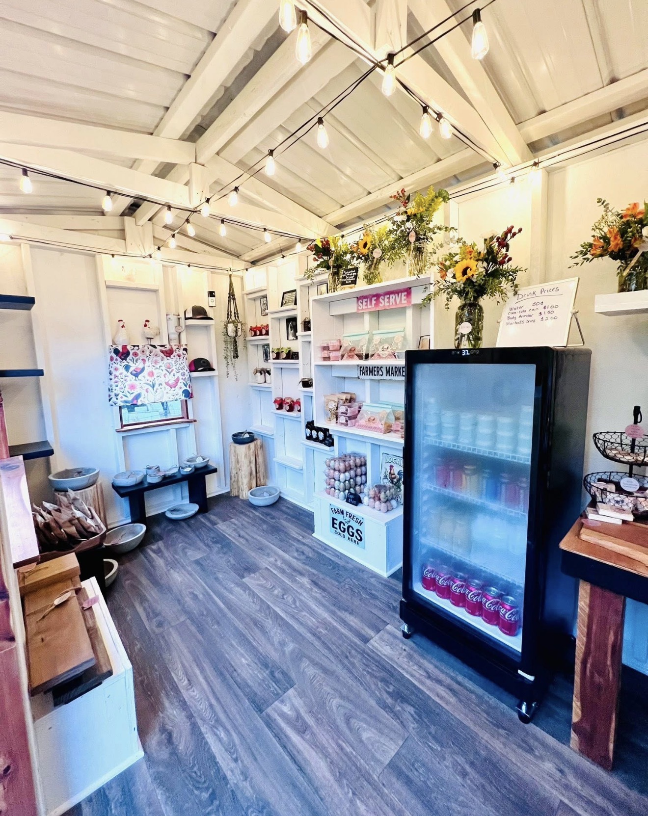 Interior of a shop with white walls, shelves, a refrigerator, and floral arrangements, with wooden flooring and ceiling beams.