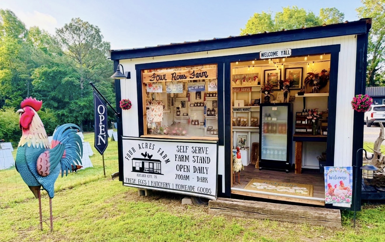 Small farm stand with open door, signs, and a display case, surrounded by greenery and decorative rooster figures, sunny day.