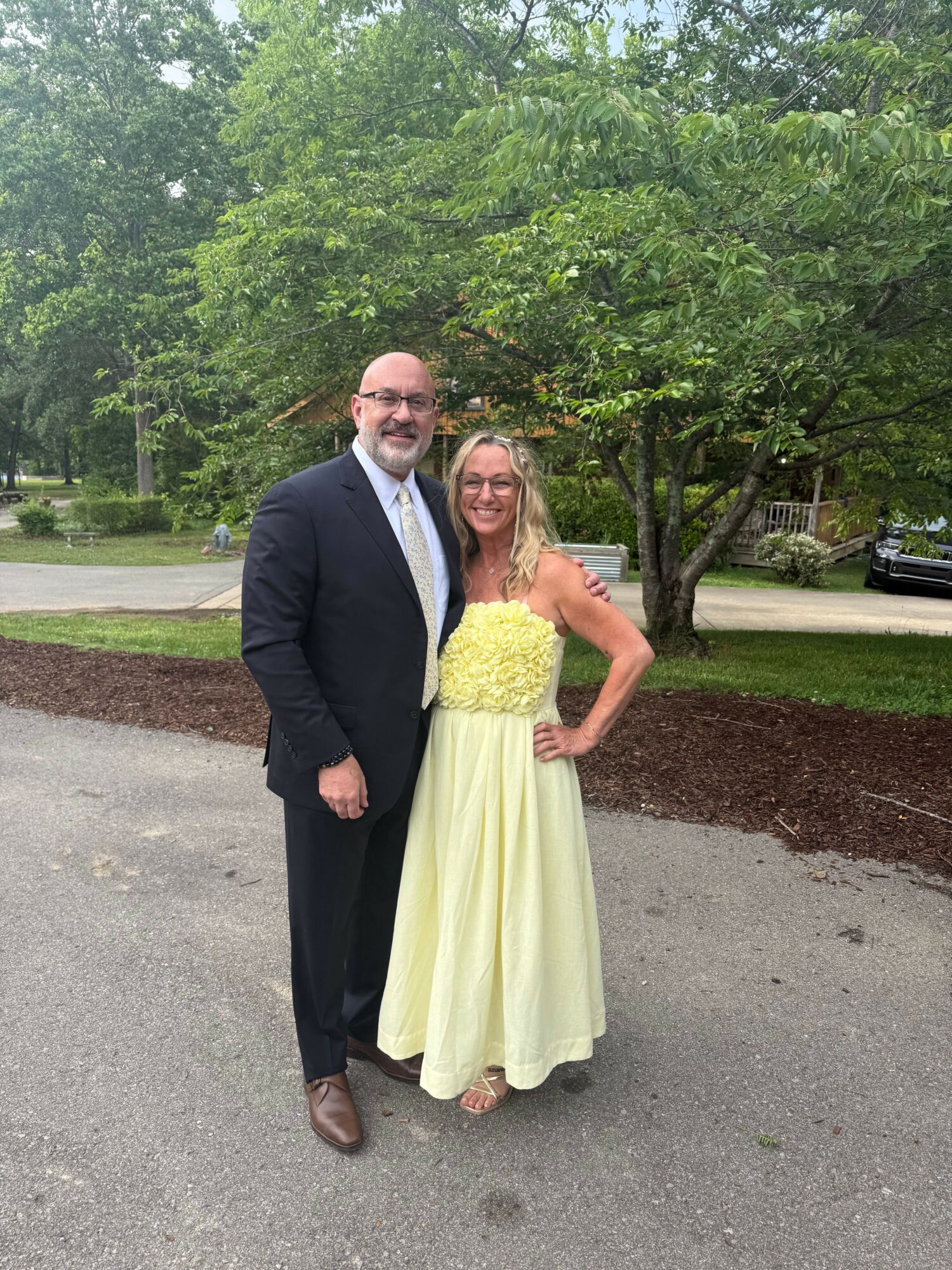 A woman in a yellow dress and a man in a suit stand outdoors on a paved path with green trees in the background.