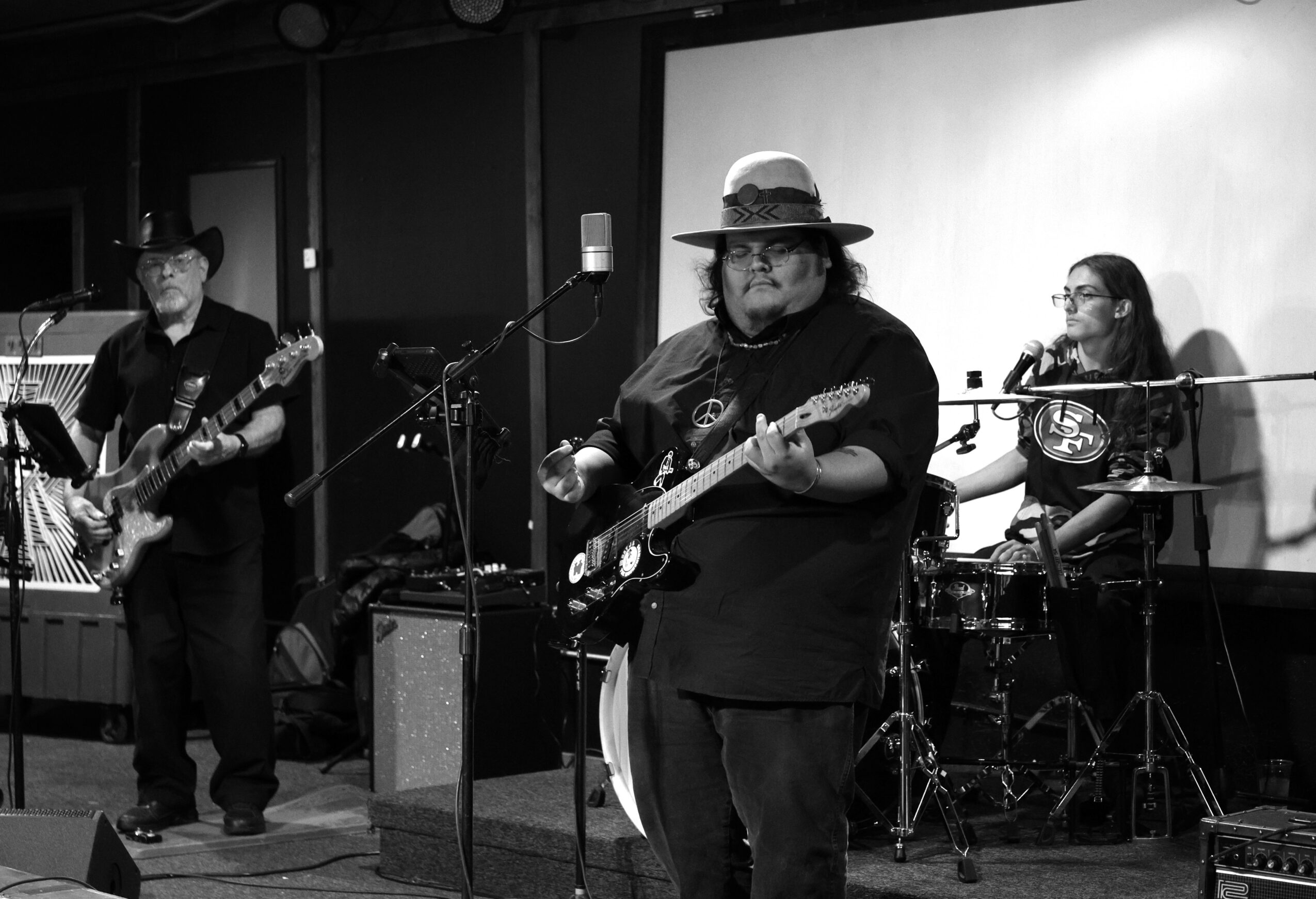 Three musicians perform on stage with guitars and drums, one wearing a hat, in a black-and-white photo.