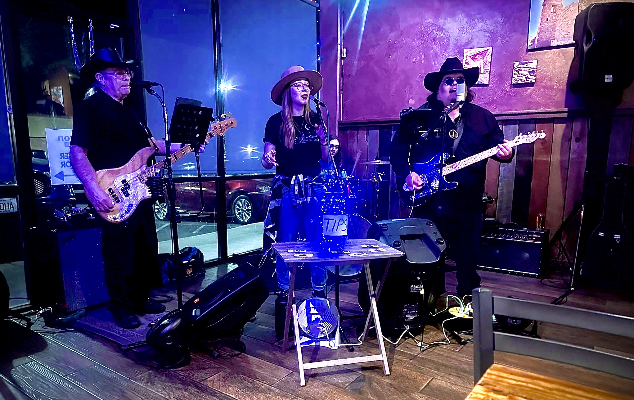 Three musicians perform on stage with guitars and a drum set, illuminated by colorful stage lights.