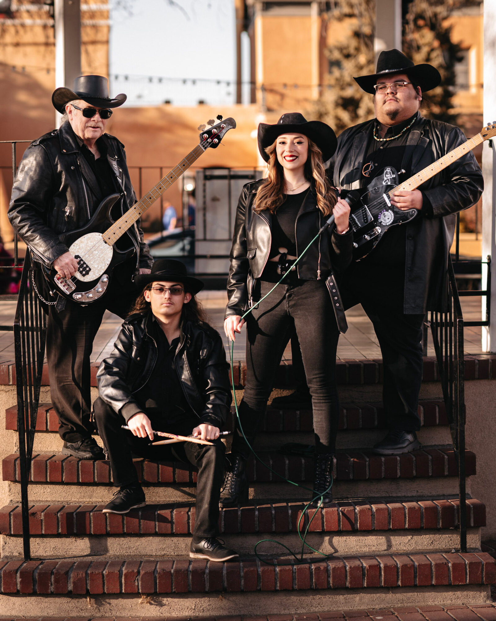 Four musicians in black leather outfits and cowboy hats pose with instruments outdoors on brick steps.