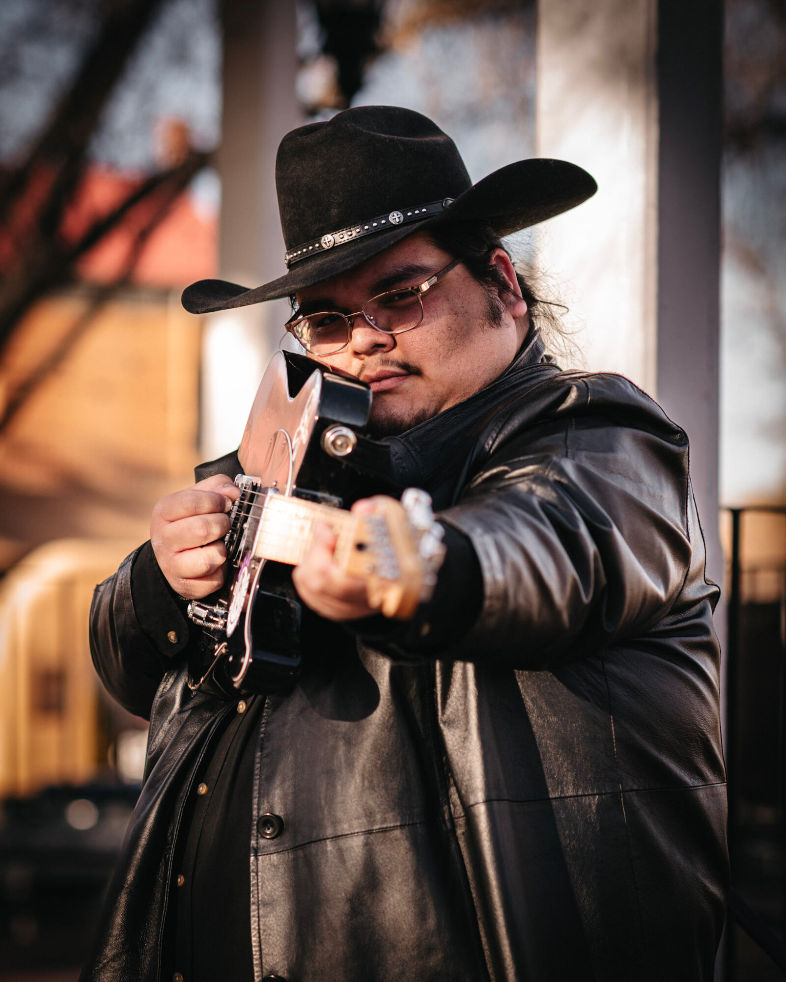 Person wearing a black cowboy hat and sunglasses, aiming a gun outdoors.