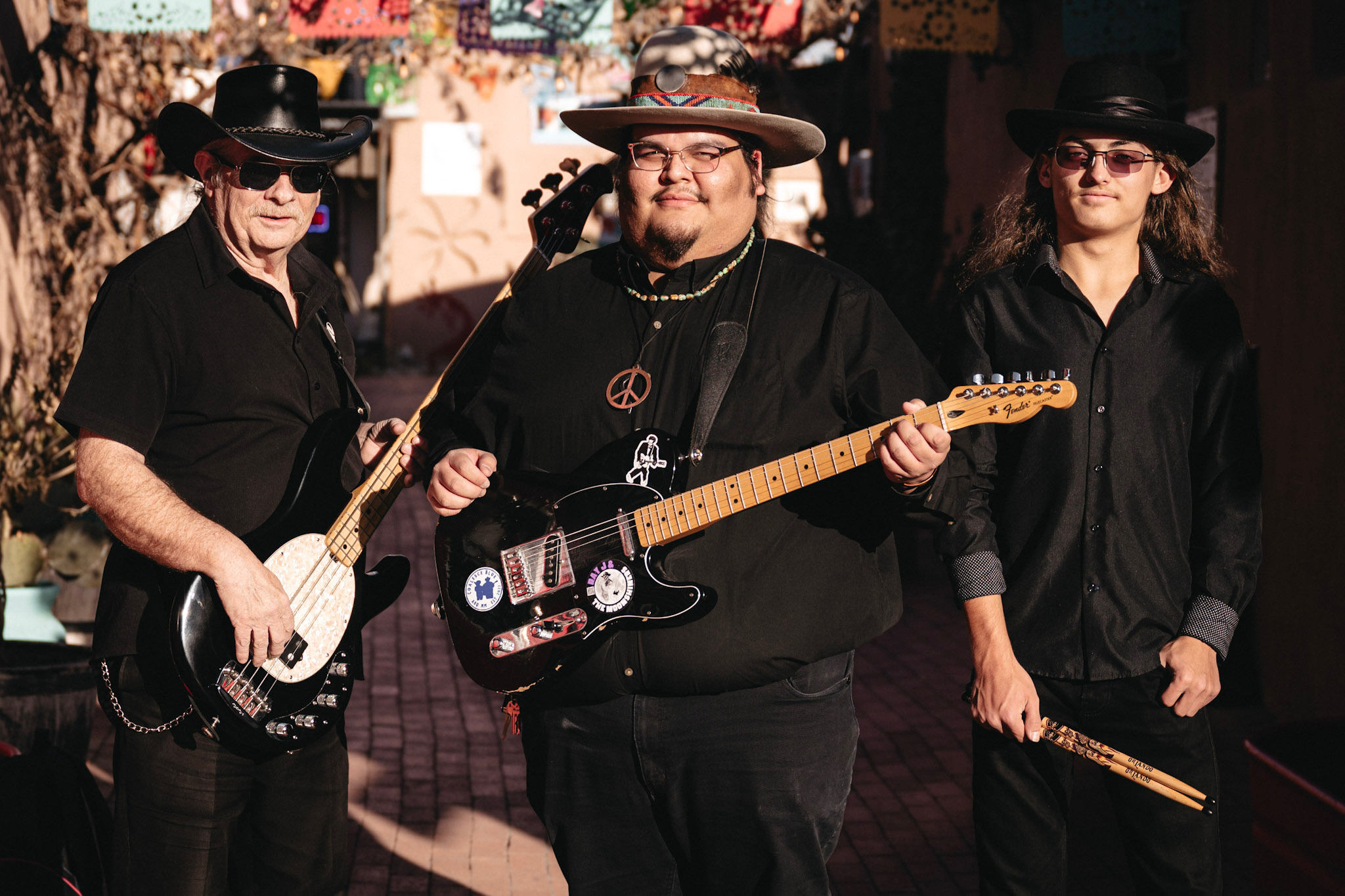 Three musicians wearing black clothing and hats, holding guitars and drumsticks, standing outdoors with trees and buildings in background.