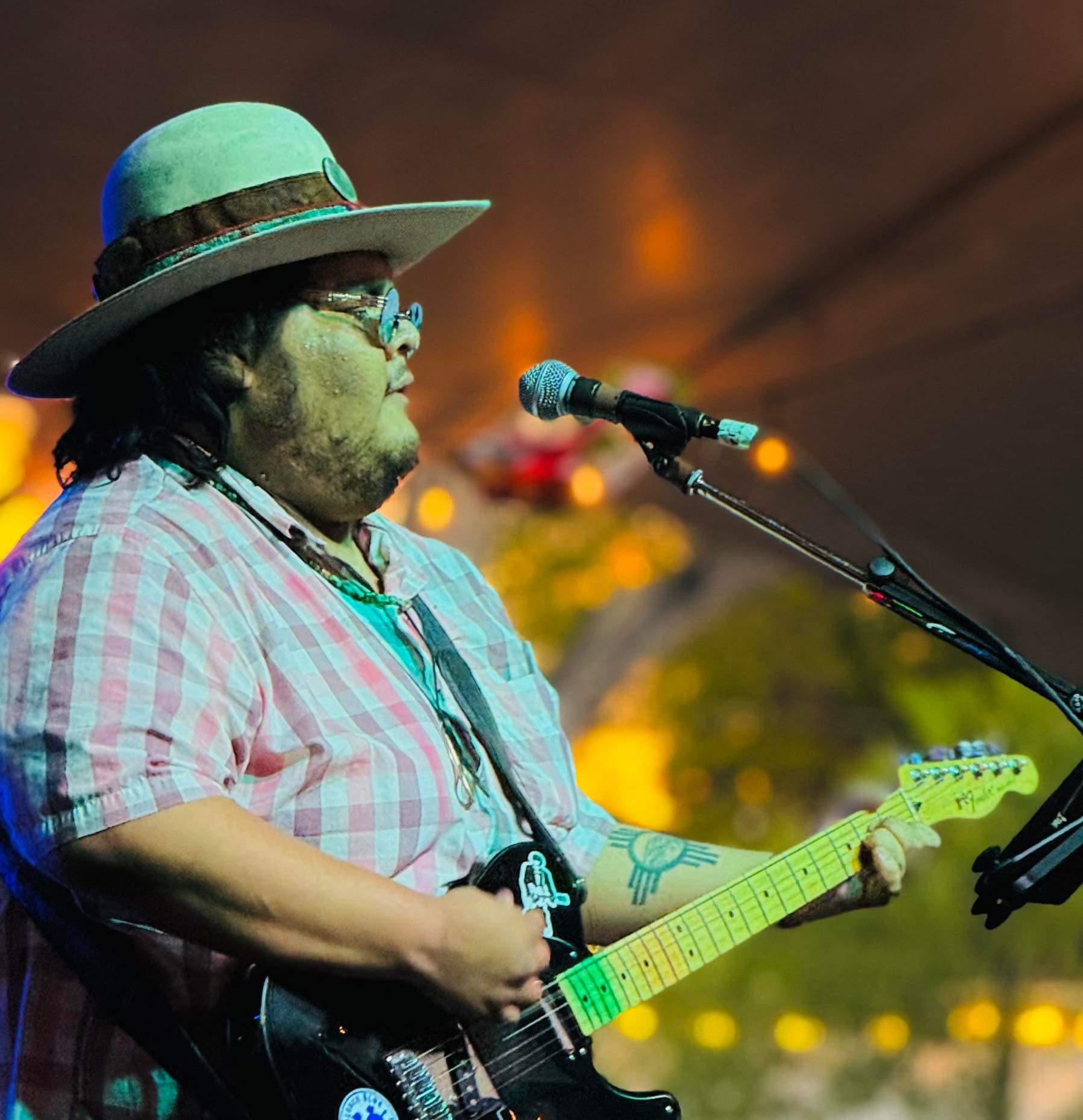 Person wearing a hat and glasses playing an electric guitar on stage with a microphone nearby.