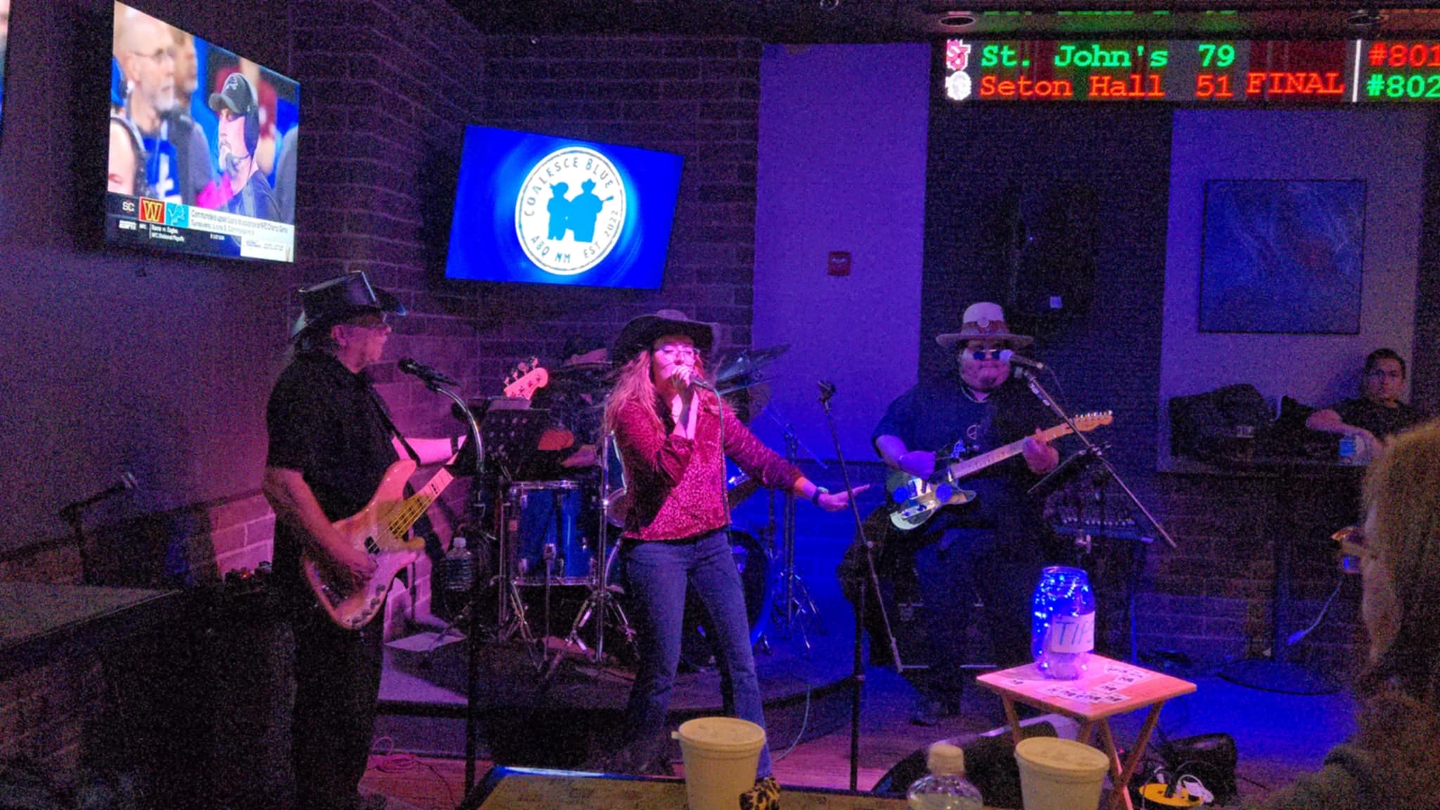 Band performs live music on stage with three musicians, a singer, and a drummer, in a dimly lit venue.
