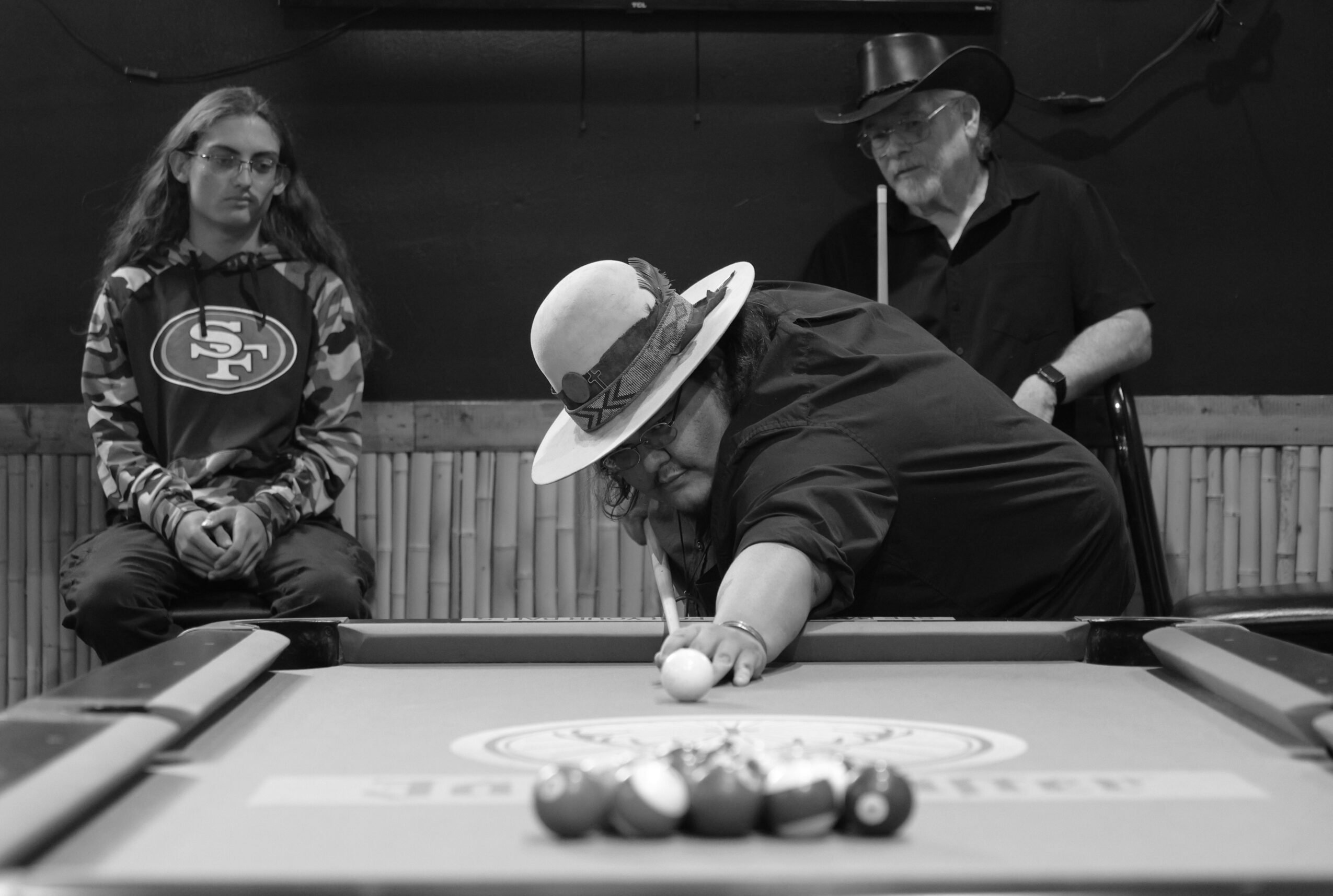 Person in a hat takes a shot at a pool table, two others watch, in a dimly lit room.