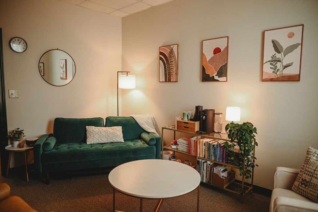 Living room with green sofa, round white coffee table, bookshelf, wall art, and lamps, arranged in a cozy setting.