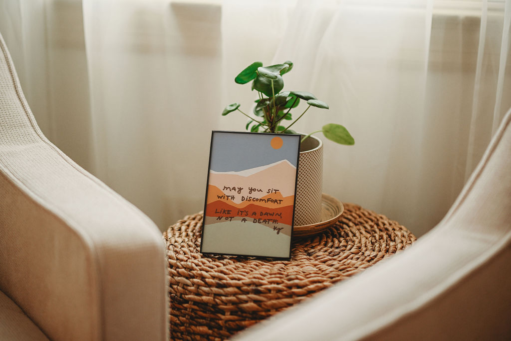 A framed picture, a potted plant, and a small object on a woven table between two beige chairs, with a sheer curtain background.