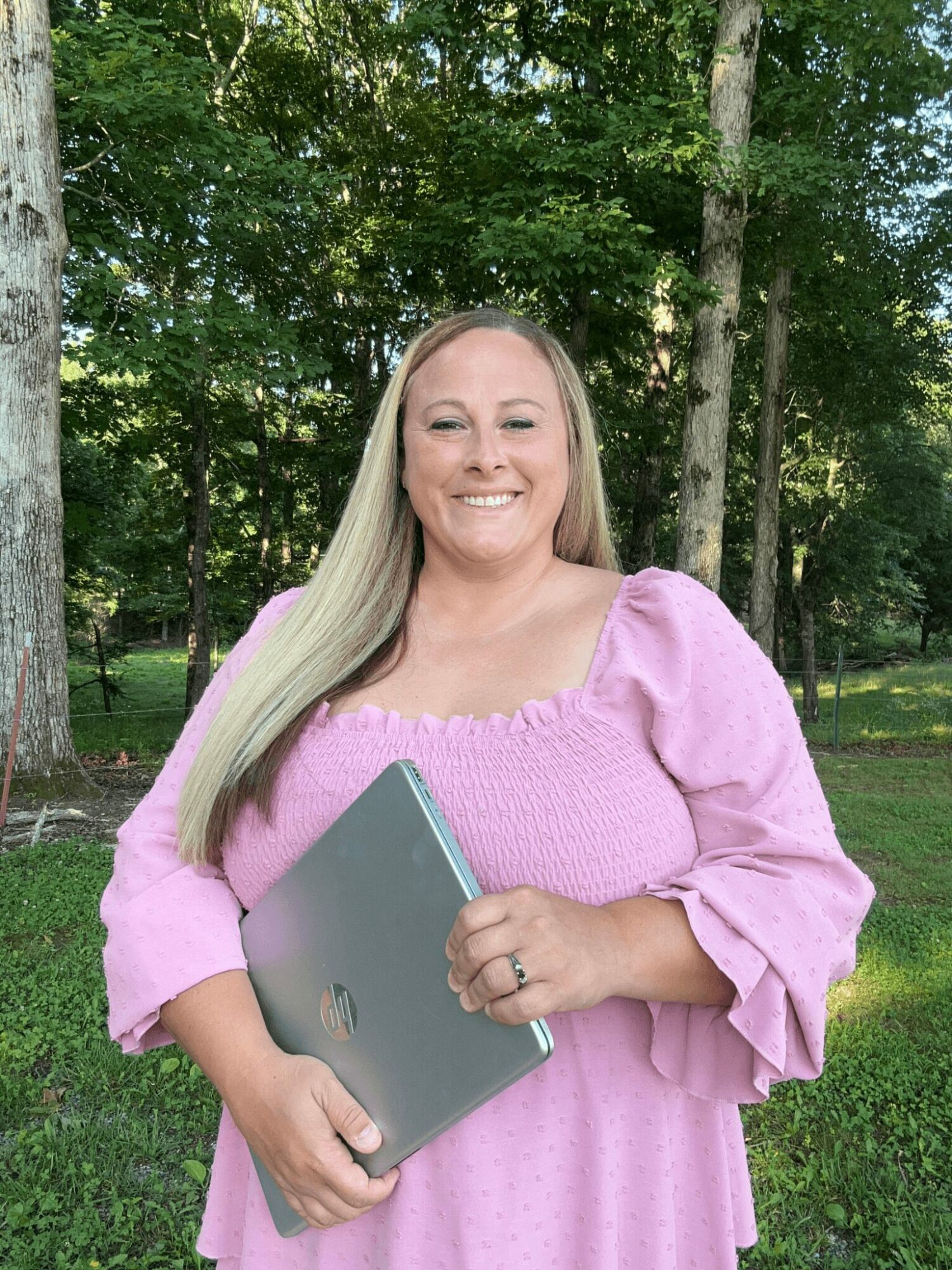 Smiling woman with long blonde hair holding a laptop outdoors in a park with trees and grass.