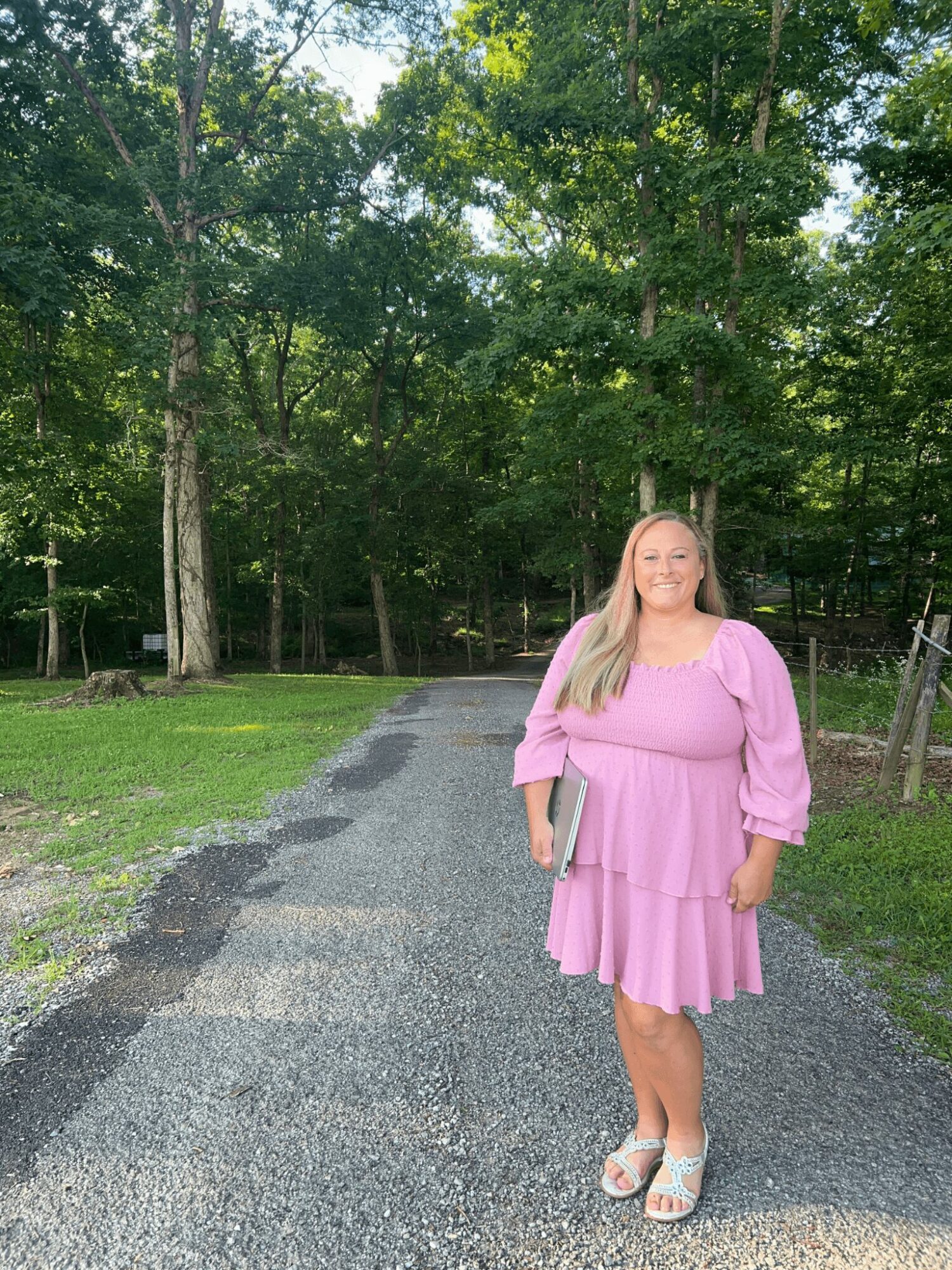 Woman in pink dress standing on a gravel path surrounded by green trees, holding a book, smiling.