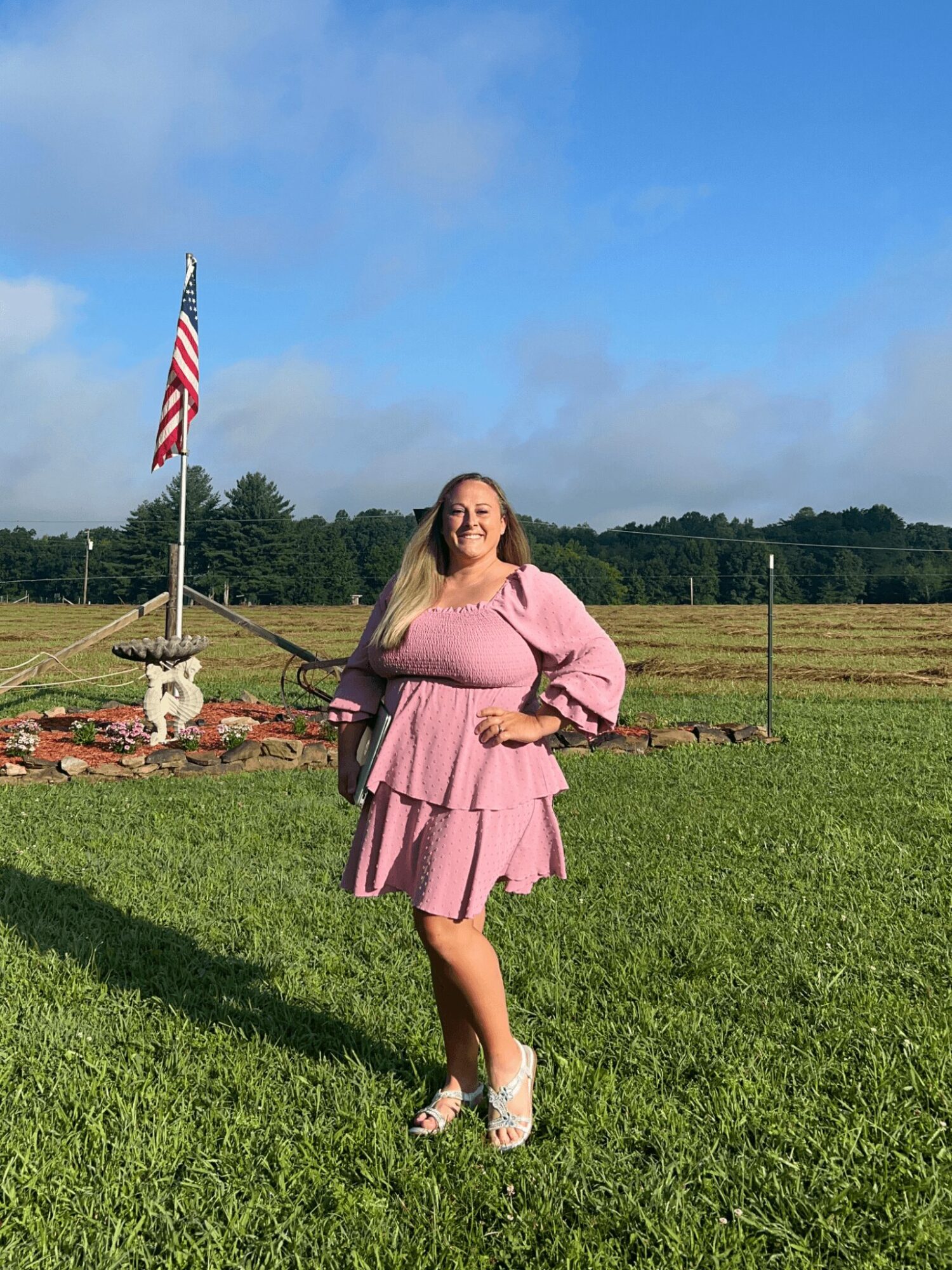 Woman in pink dress standing outdoors on grass with American flag and blue sky background.