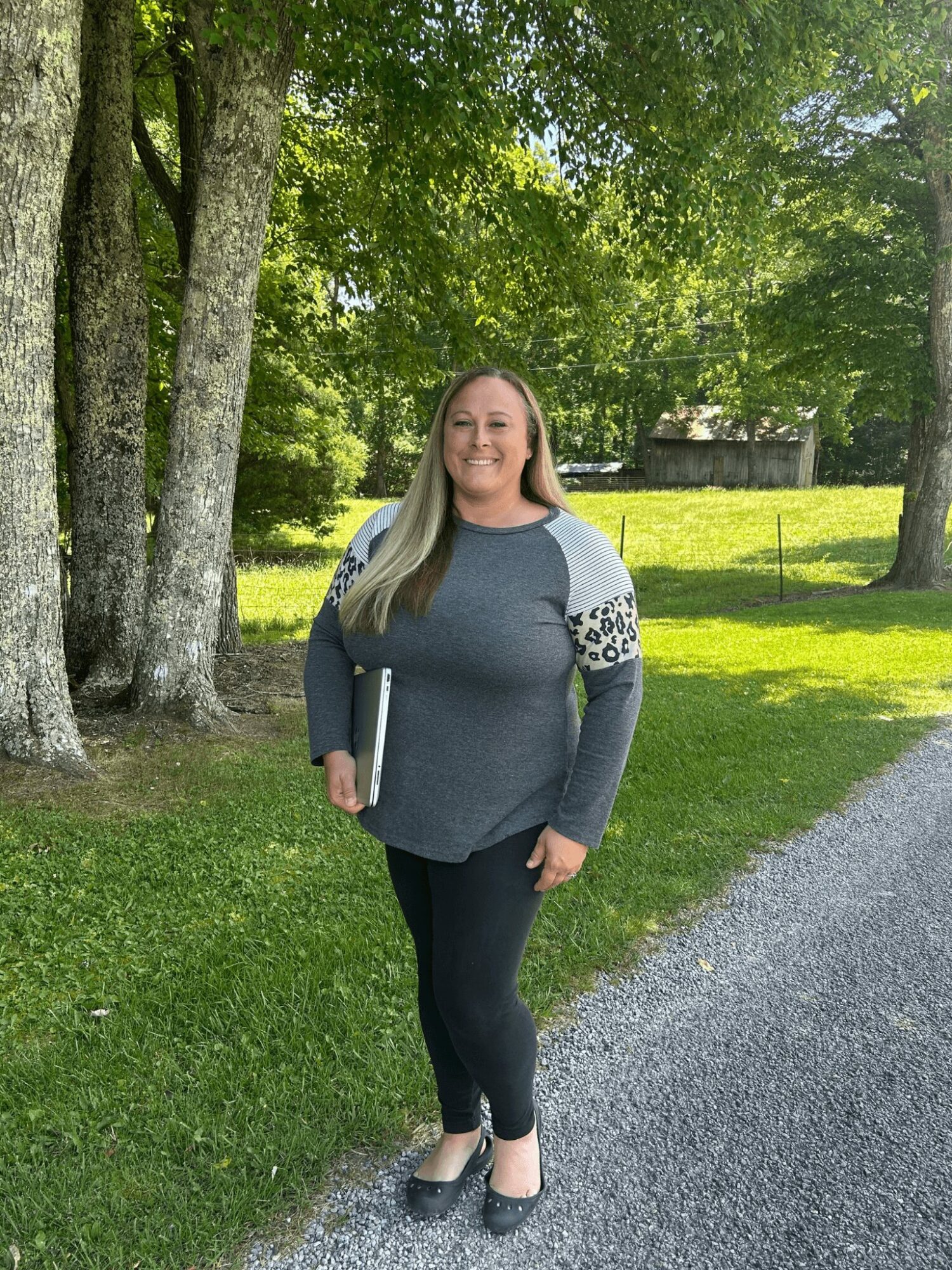 Woman standing outdoors on a gravel path, holding a book, smiling, with trees and grass in the background.