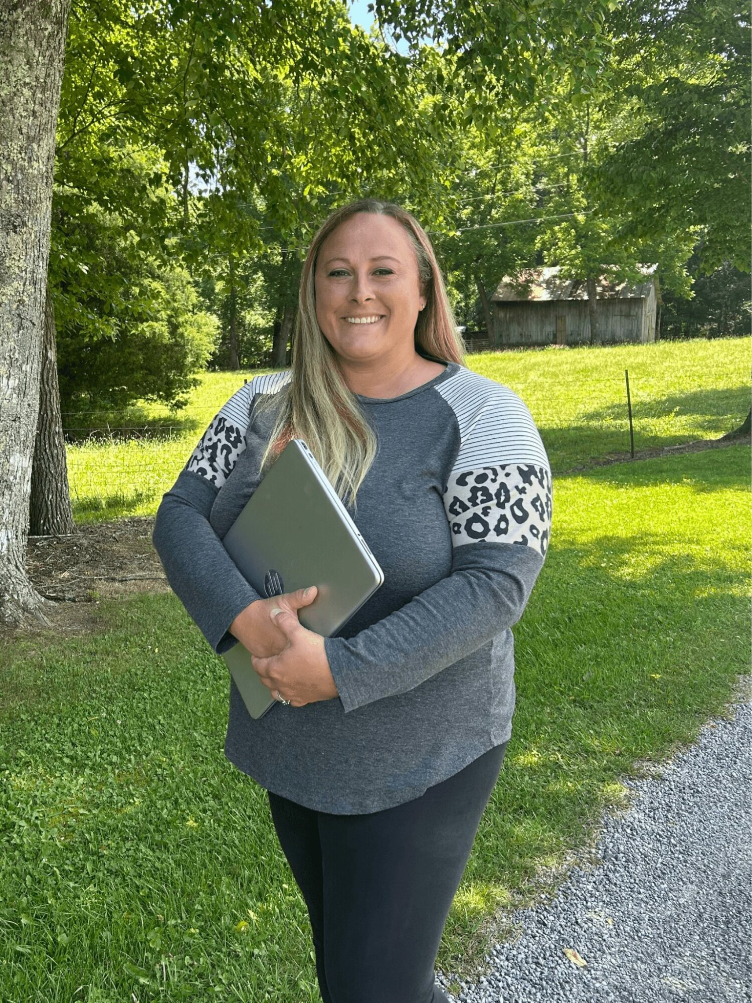 Smiling woman holding a laptop outdoors in a park with trees and grass.